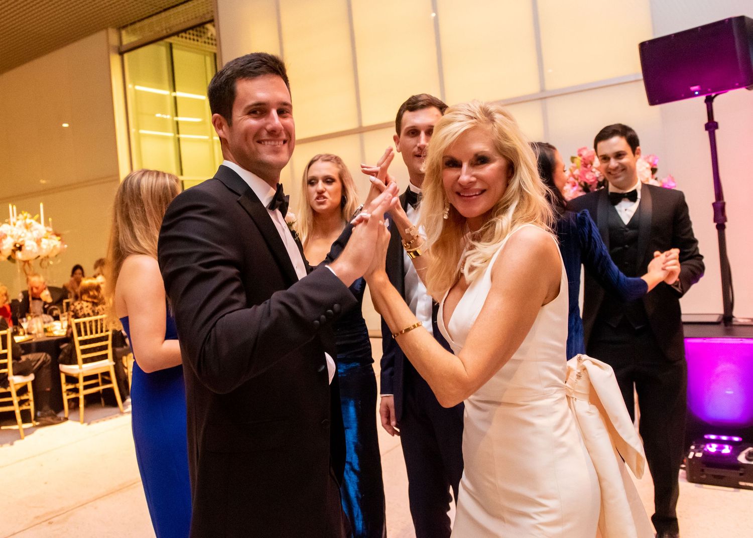 Bride and groom share a dance together smiling at wedding reception with guests celebrating behind them.