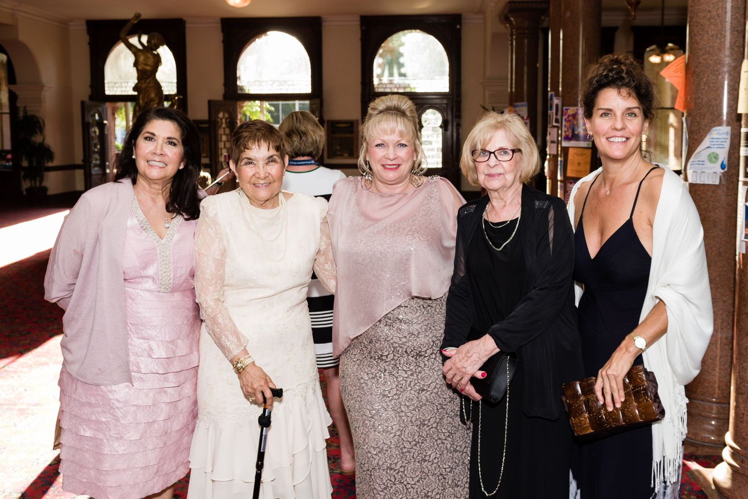 Five elegantly dressed women posing together indoors at a formal event, wearing evening gowns and dressy attire.