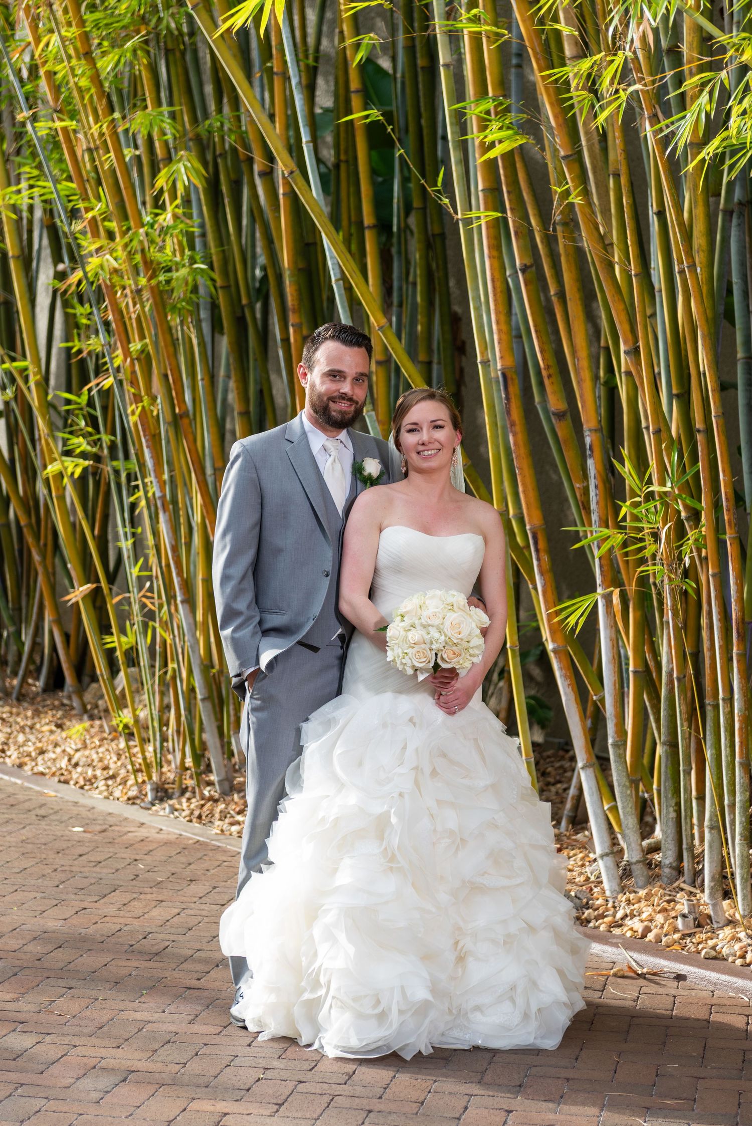Bride and groom pose together in front of lush bamboo grove, bride holding white bouquet in ruffled strapless gown.