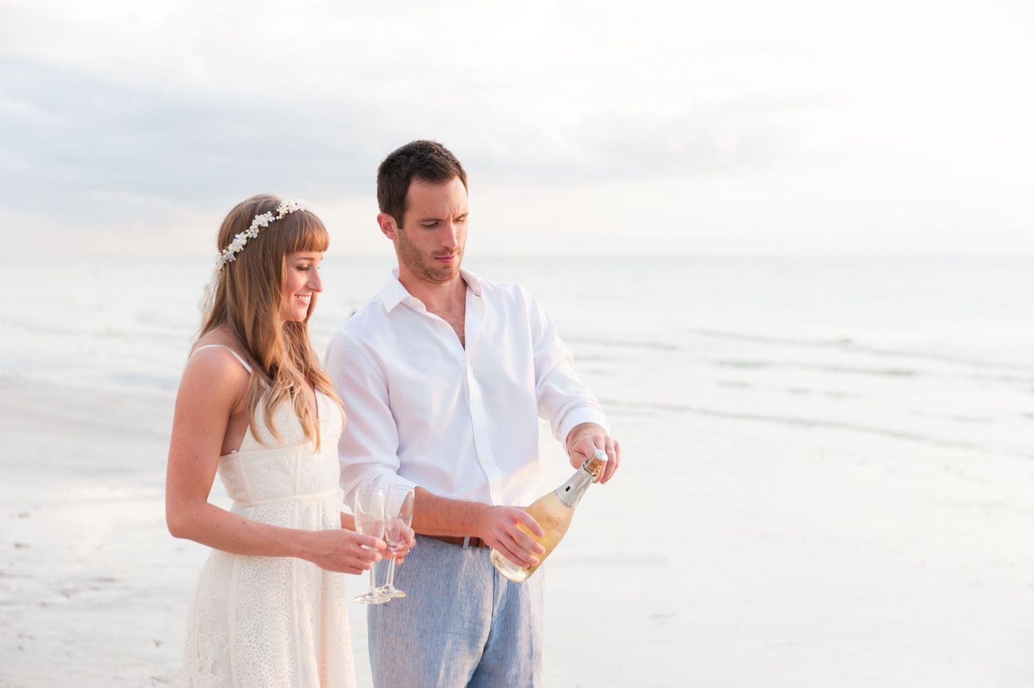 A couple in white attire celebrates on a beach, pouring champagne at sunset.