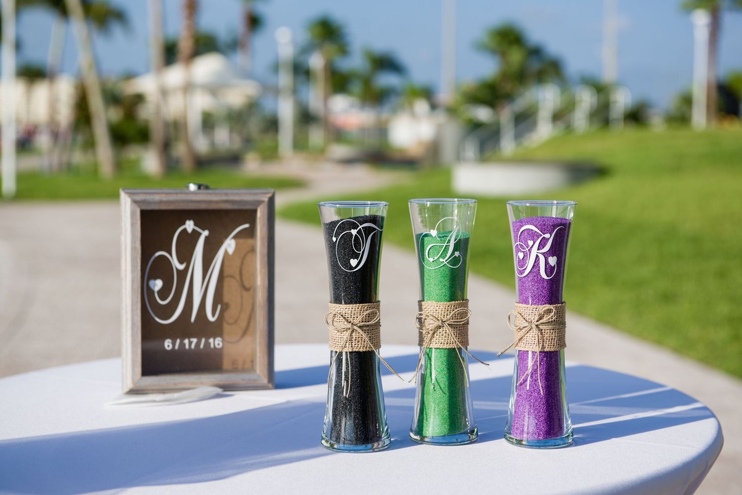 Three monogrammed sand ceremony vases in black, green, and purple with twine bows beside a framed wedding date sign.