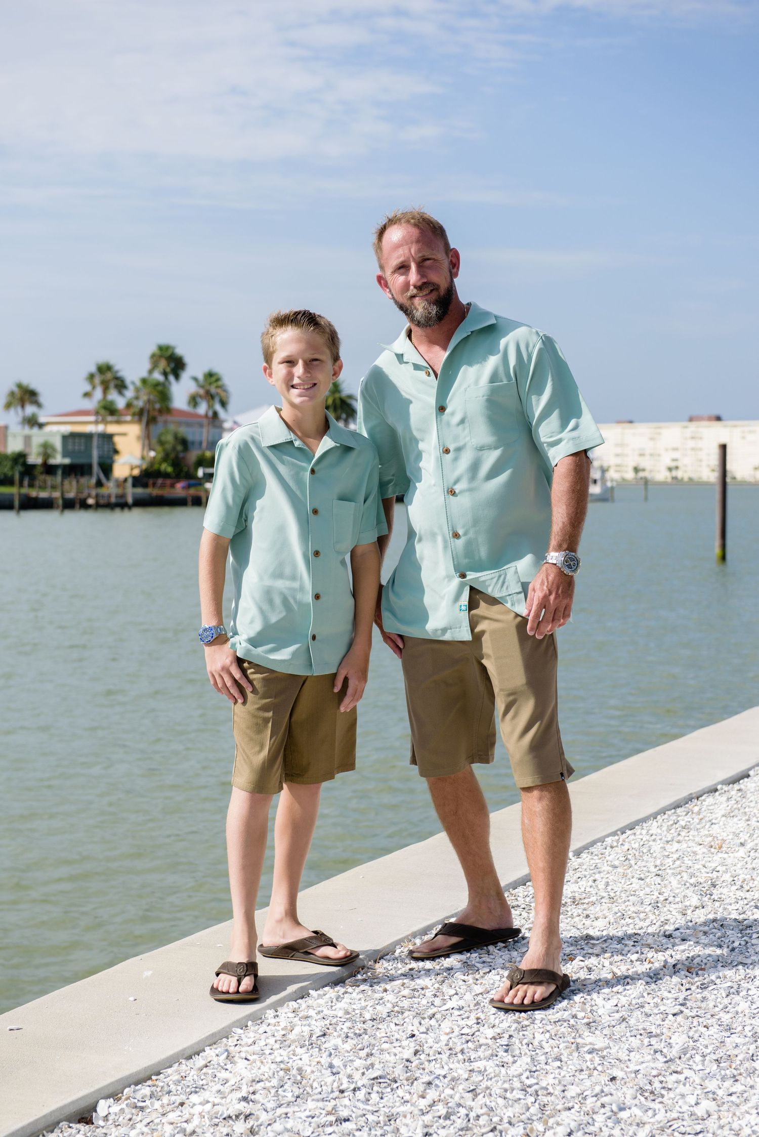 Father and son wearing matching mint green shirts and khaki shorts posing together by a waterfront on a sunny day.