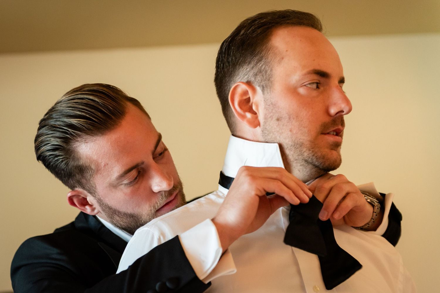 A man in a tuxedo adjusts another mans black bow tie while getting ready for a formal event.