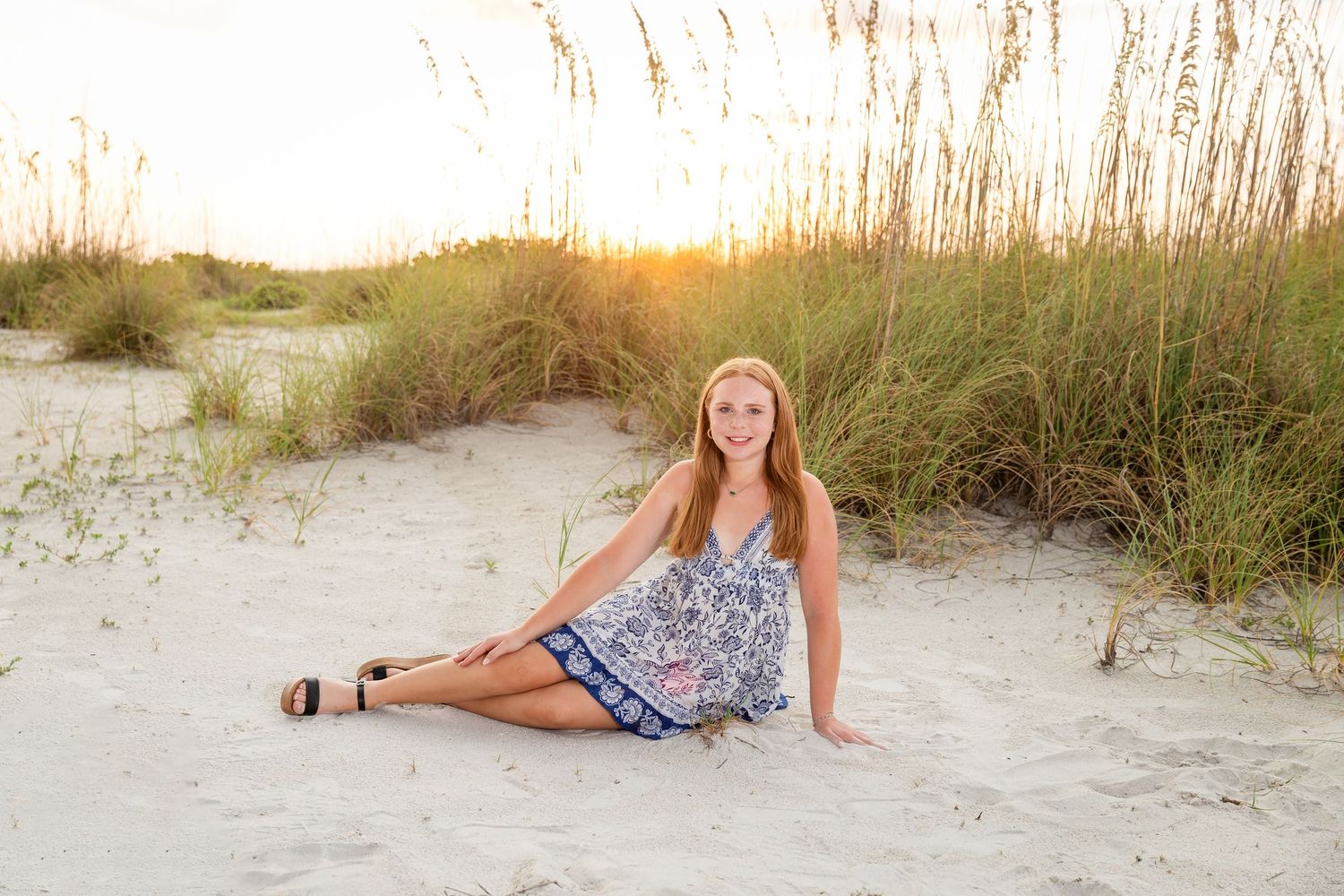 Young woman in floral dress sitting on sandy beach dunes at golden sunset with sea oats in background.