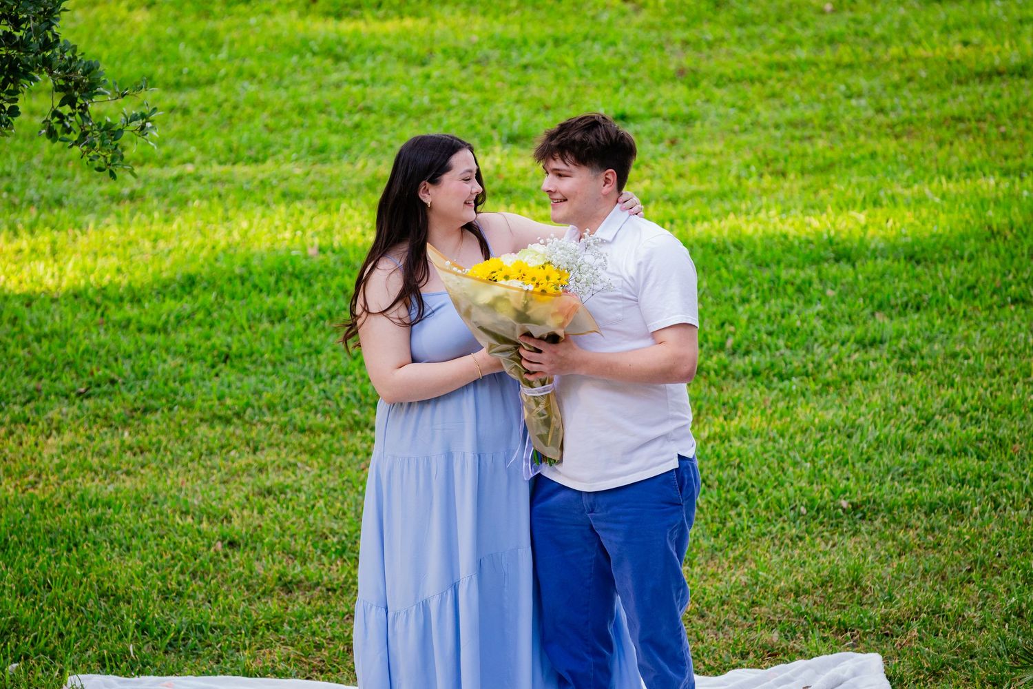 Couple sharing yellow flower bouquet on green lawn, woman in blue dress, man in white shirt and blue pants.