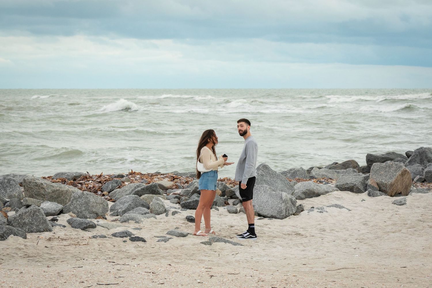 Couple sharing a romantic moment on a rocky beach with choppy ocean waves under cloudy skies.