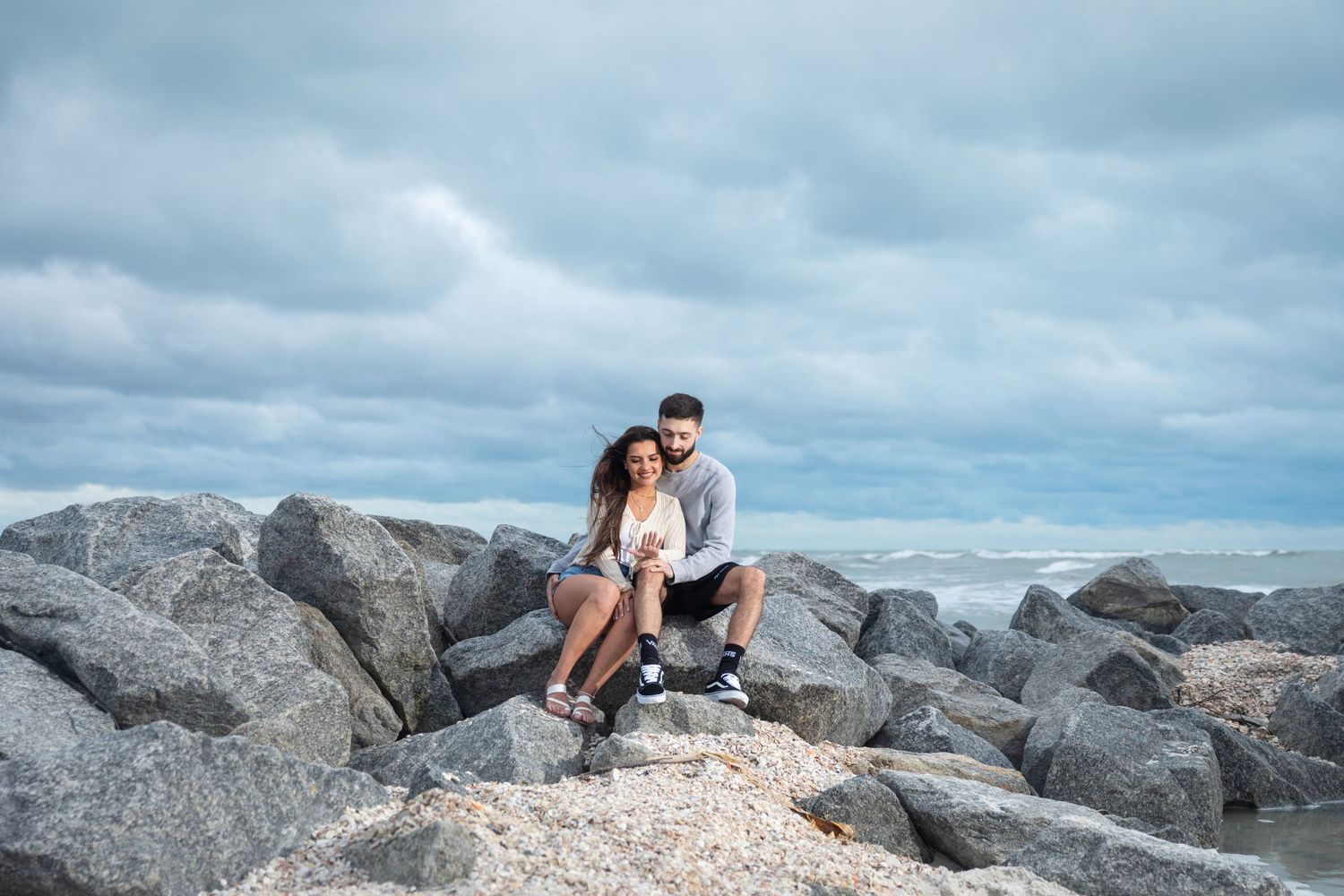 Couple sitting together on rocky shoreline with cloudy sky and ocean waves in the background.