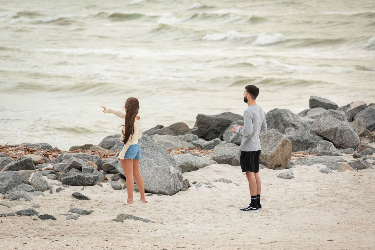 A girl and boy stand on a sandy beach near rocks, as she points toward the ocean waves.