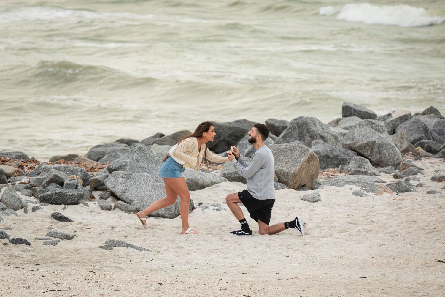 Man proposes on bended knee to woman on sandy beach with rocky shoreline and ocean waves in background.
