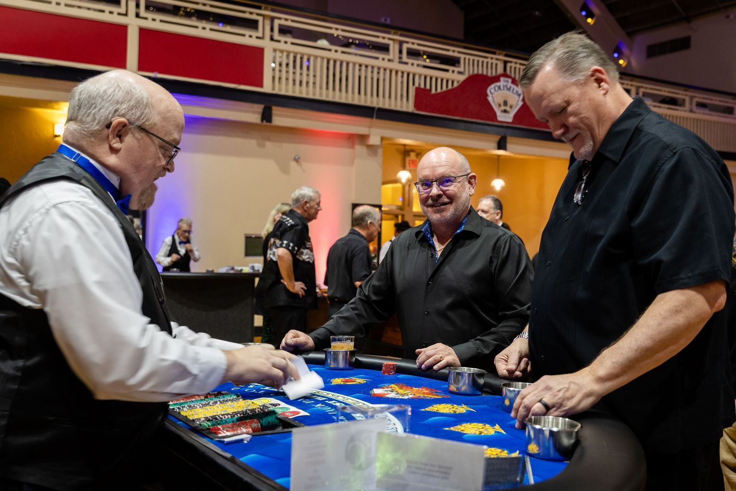 Three men enjoying a casino card game at a blue felt table during a formal event in a decorated venue.