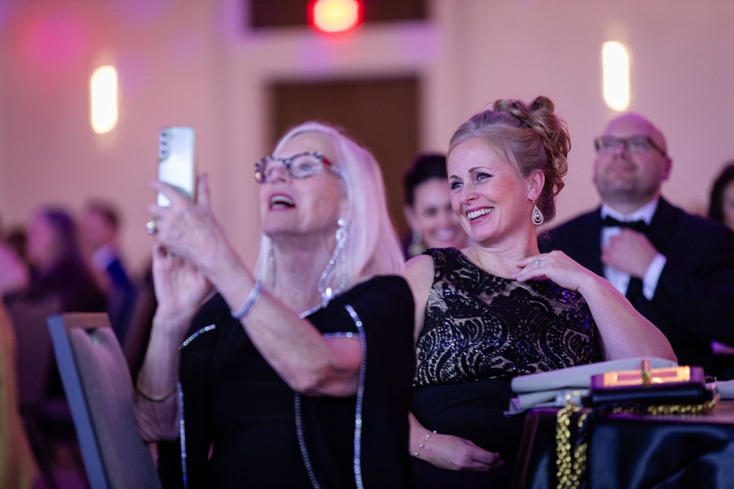 Two women in elegant black dresses laughing and taking selfie at formal gala event.