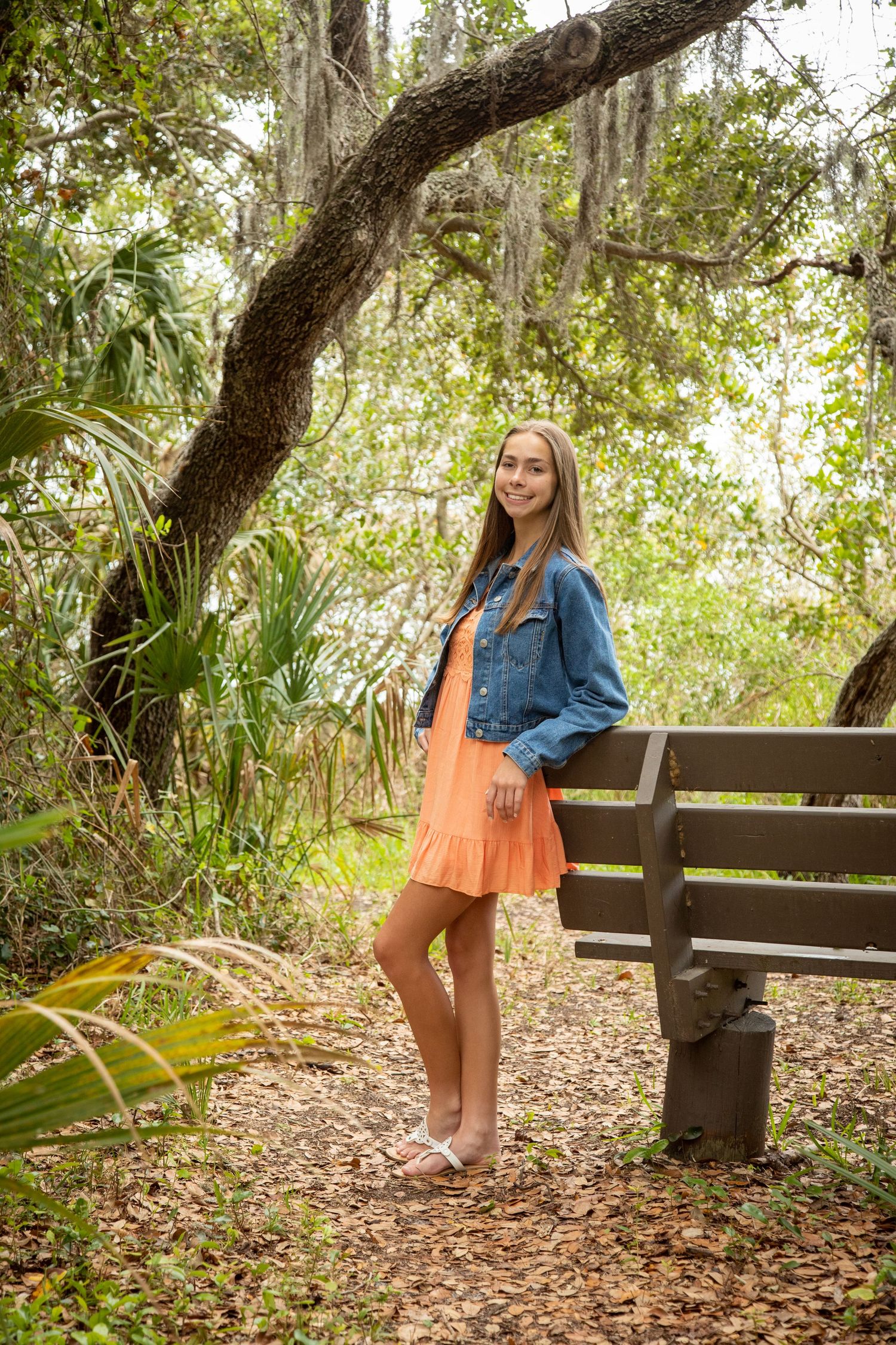 Young woman in orange dress and denim jacket smiling beside a bench on a scenic wooded nature trail.