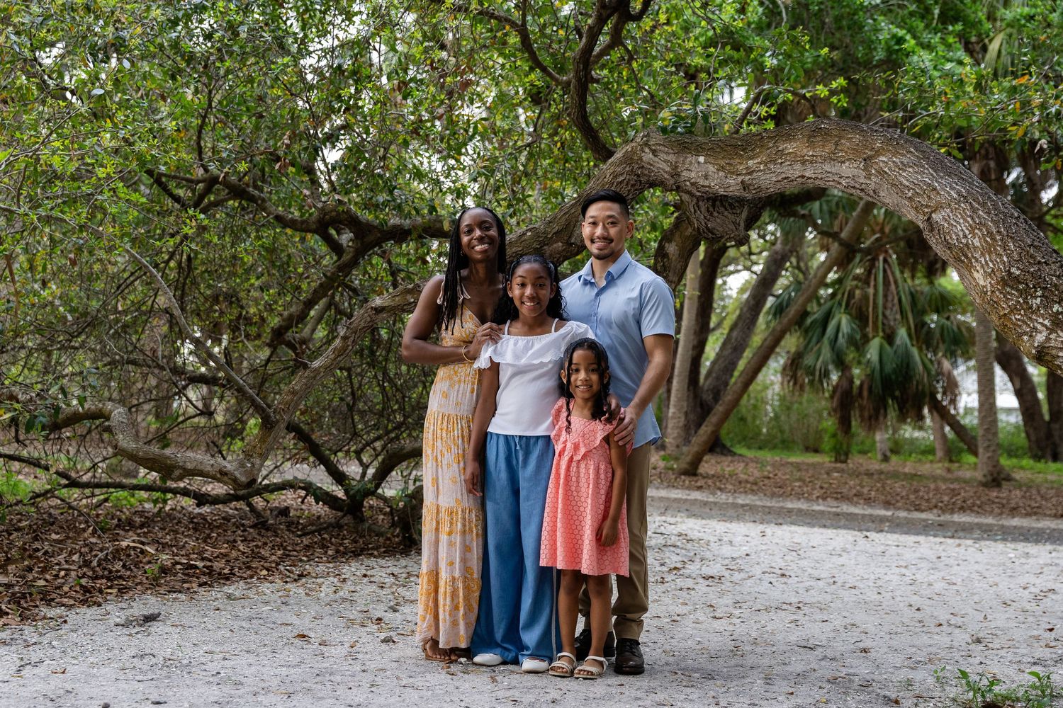 Family of four smiling together outdoors under a large tree on a sandy path in a lush green park.
