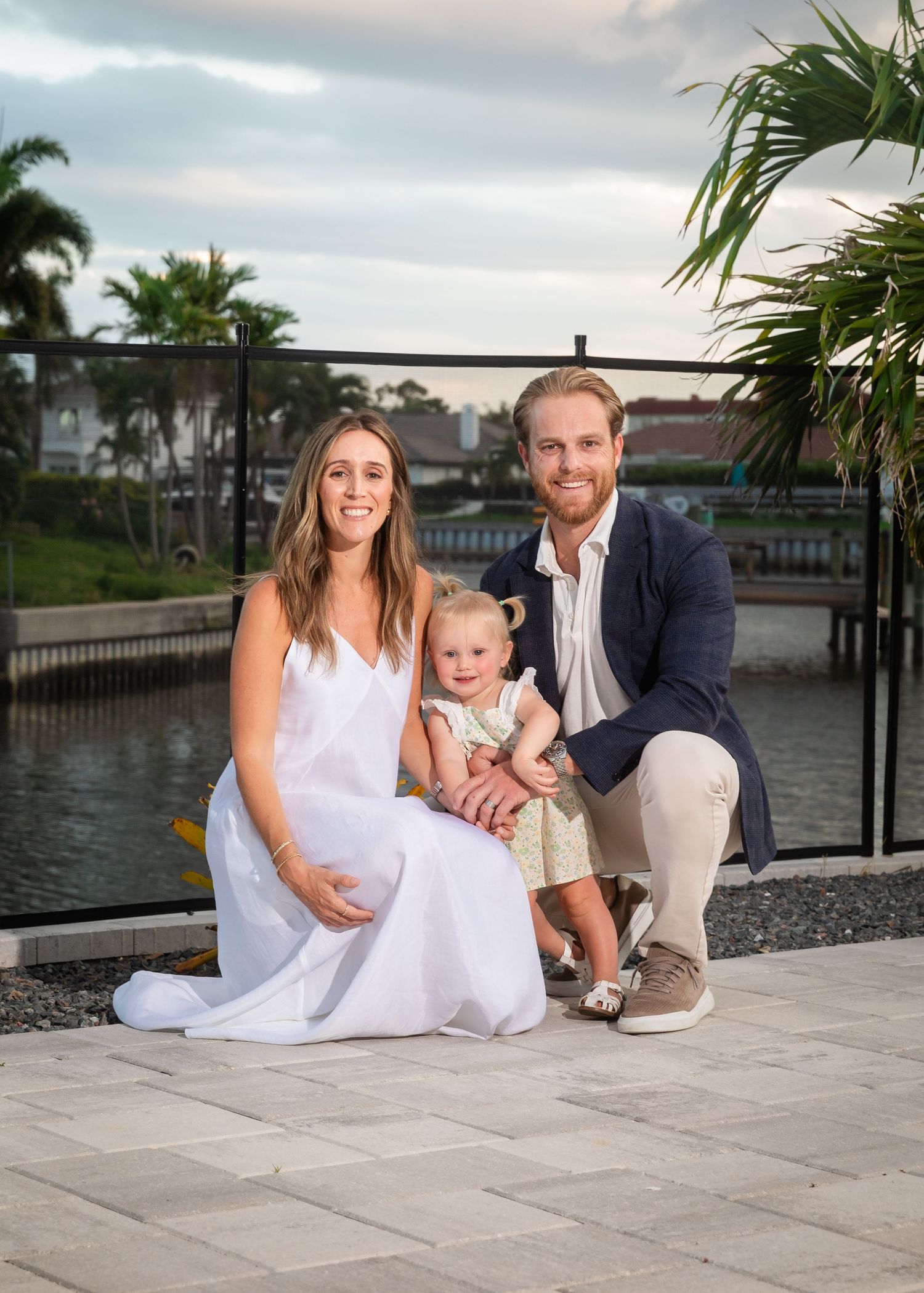 A smiling family of three poses outdoors near a waterway, with a woman in white, a man in navy, and a toddler in a floral dress.