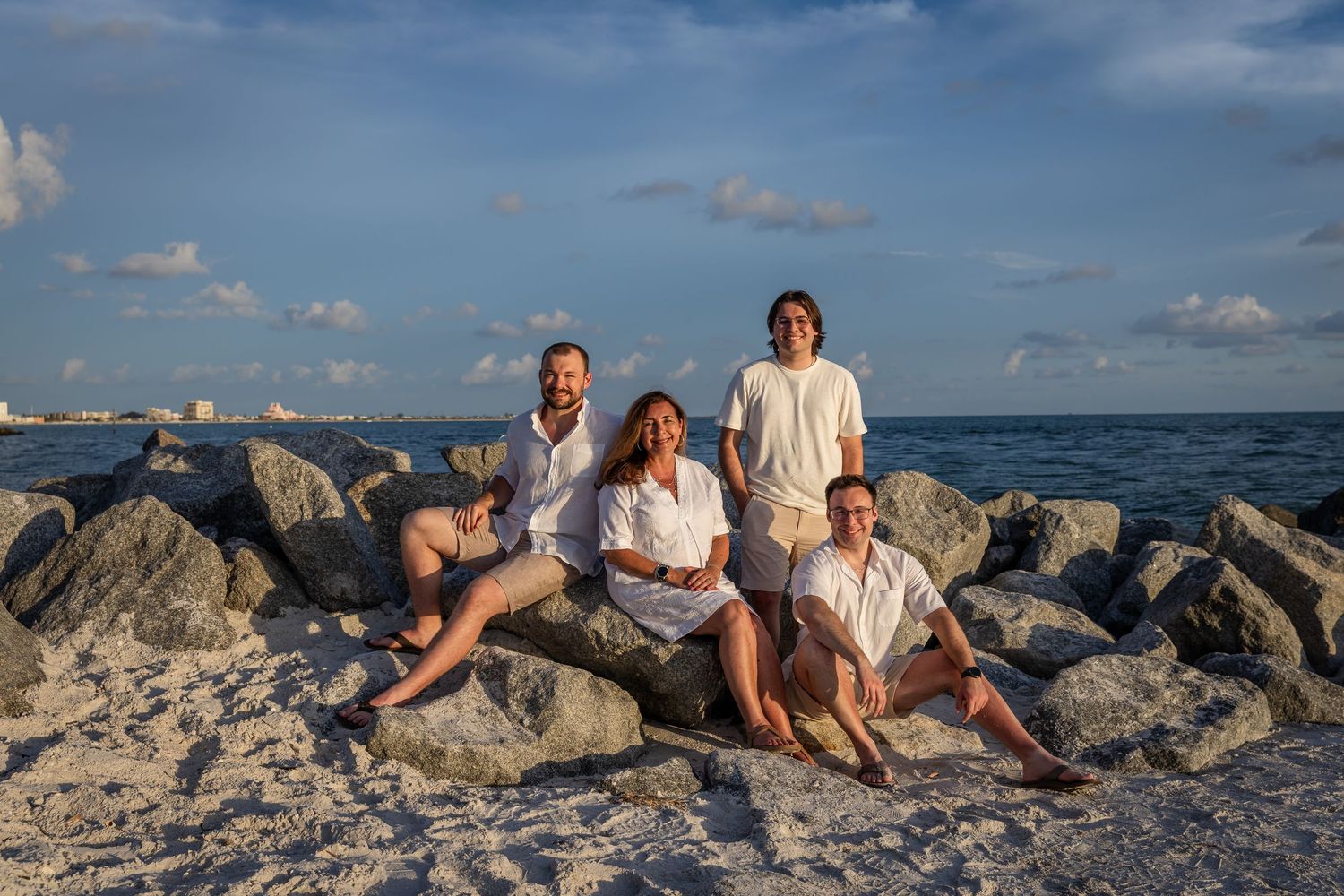 Family of four posing on rocky beach shoreline at sunset, wearing white and khaki outfits with ocean backdrop.