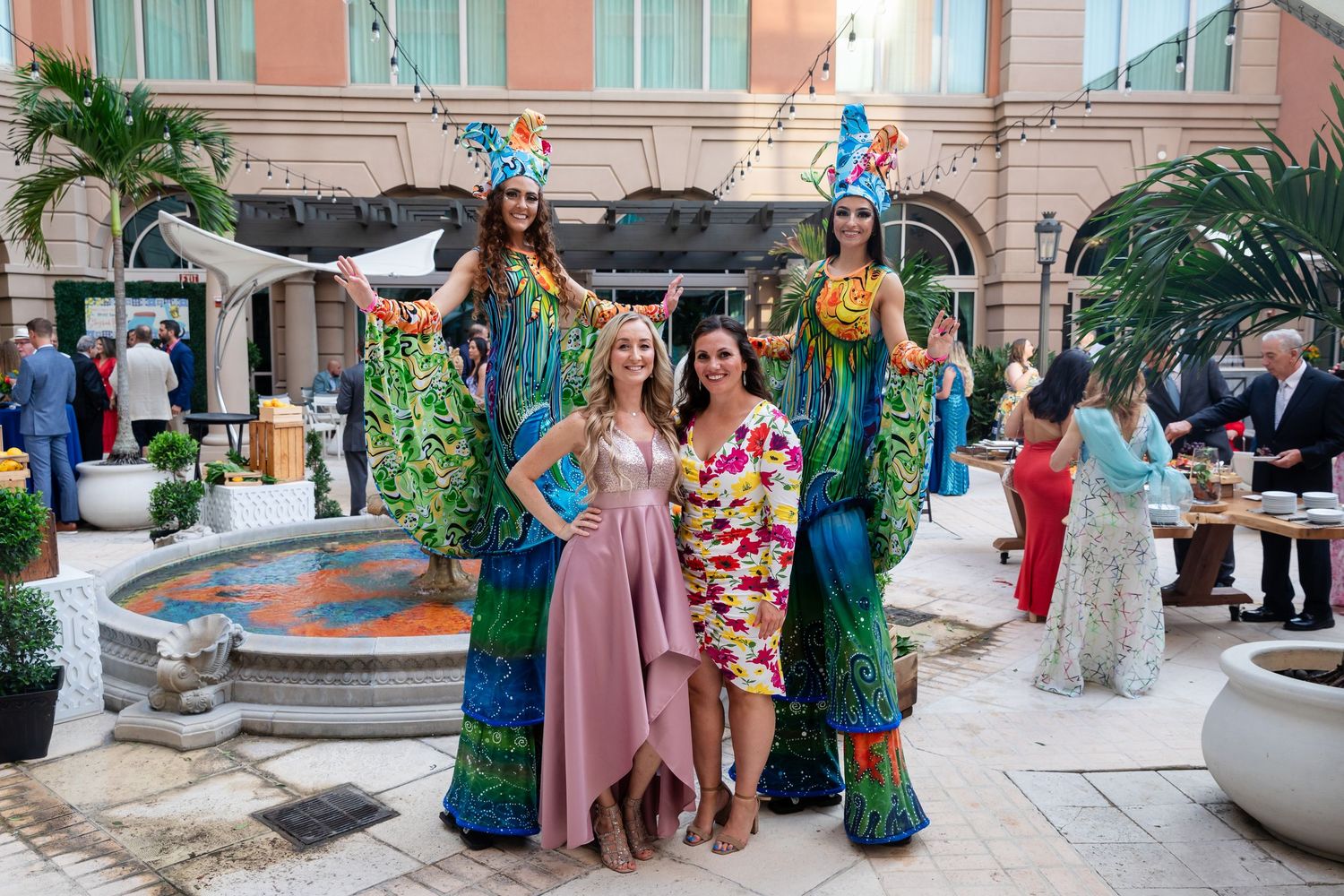 Two women pose with colorful stilt performers in tropical costumes at an outdoor luxury hotel courtyard event.