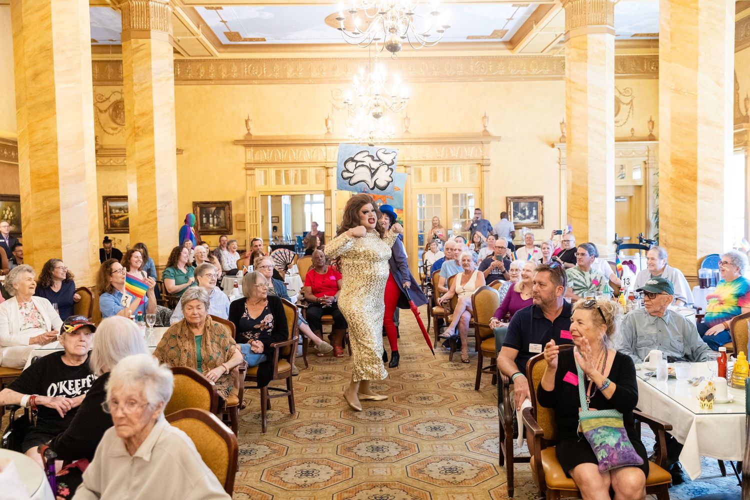 A performer in a gold costume walks a runway at an elegant banquet hall filled with seated guests during a fashion show.