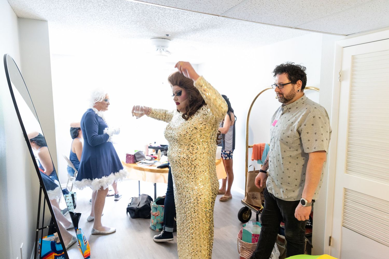 People in colorful outfits gather in a bright room, with a woman in a gold dress adjusting her hair.