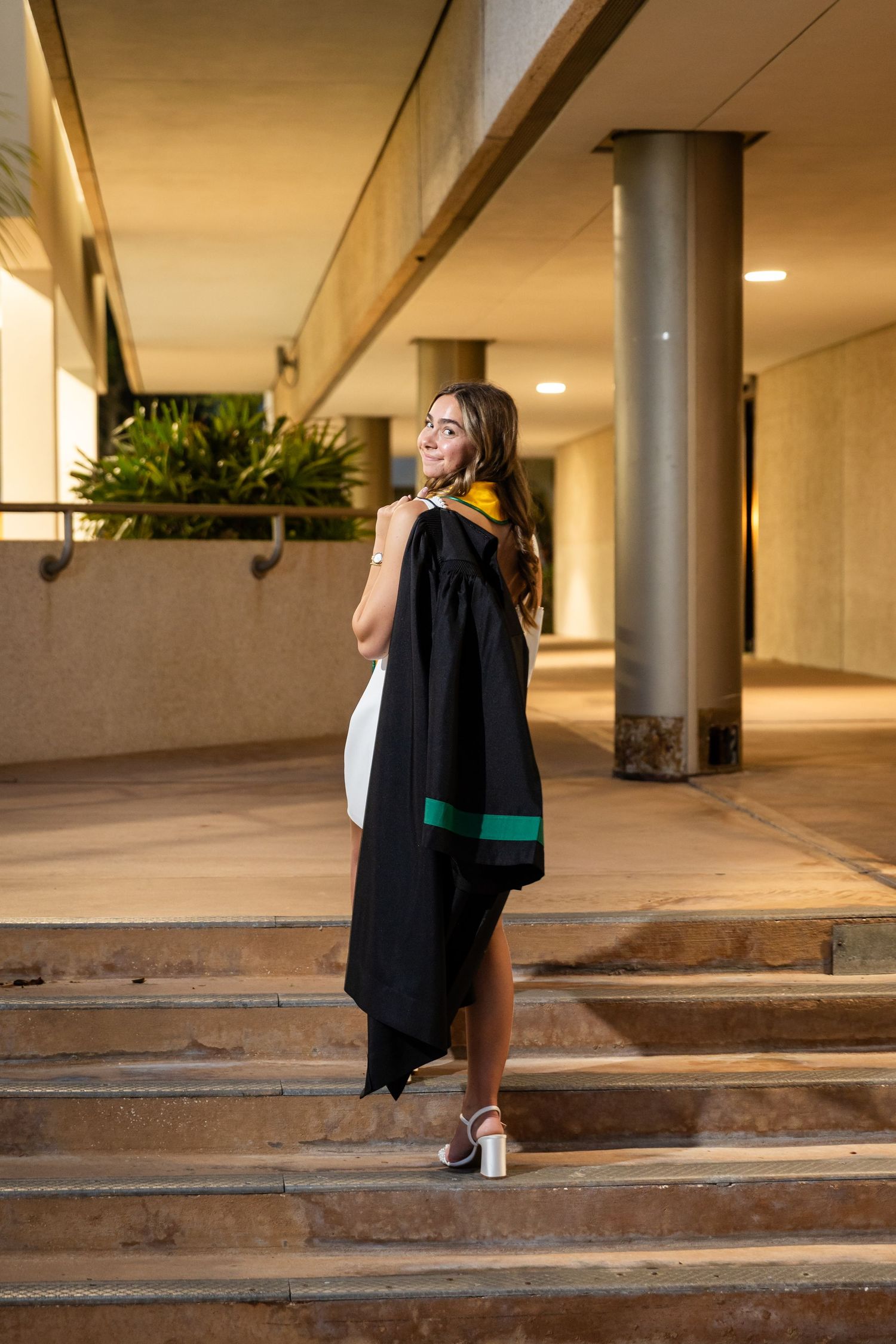 Young woman in white dress holding black graduation gown on outdoor steps, smiling over shoulder.