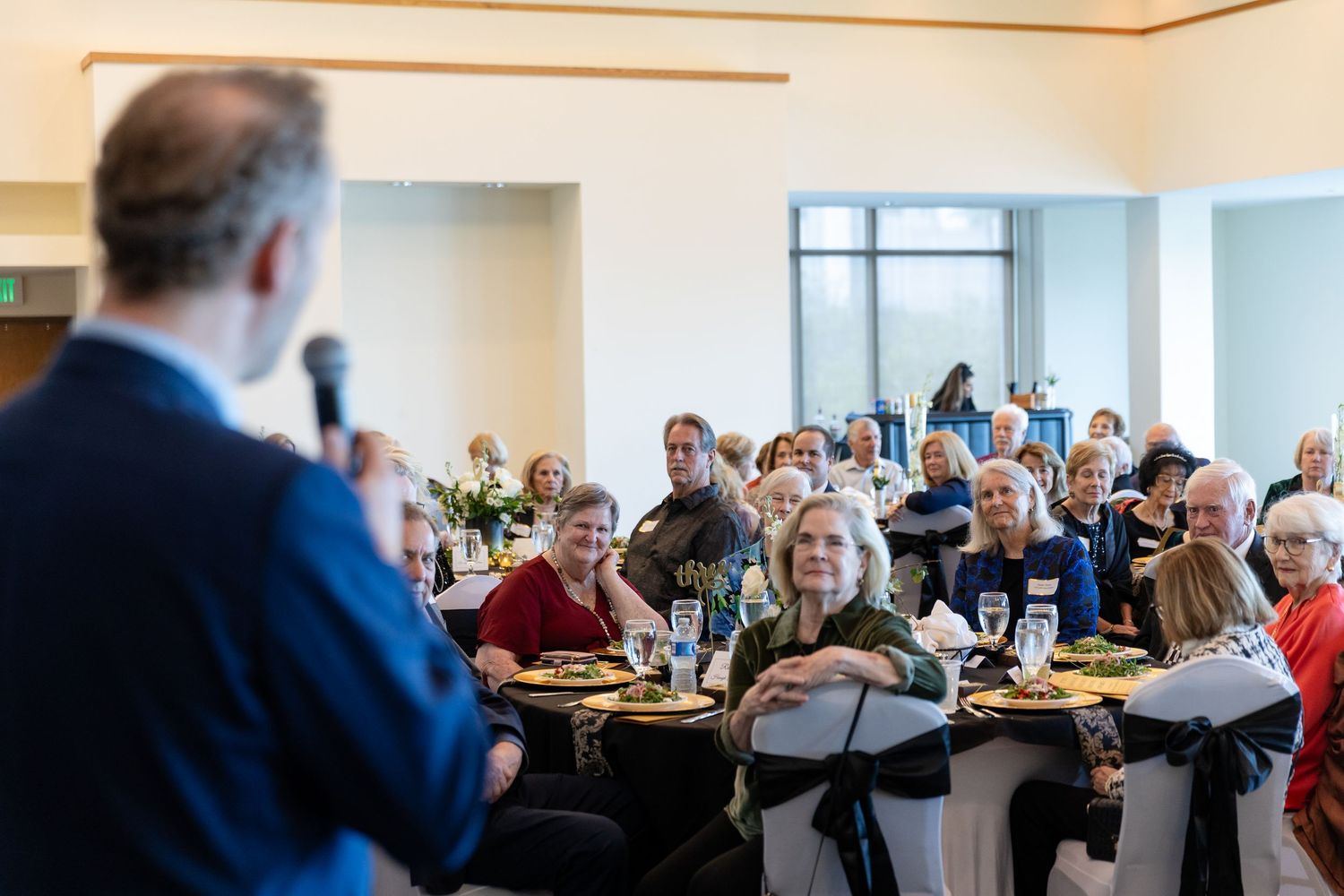 Speaker addresses large audience of guests seated at banquet tables during a formal luncheon event.