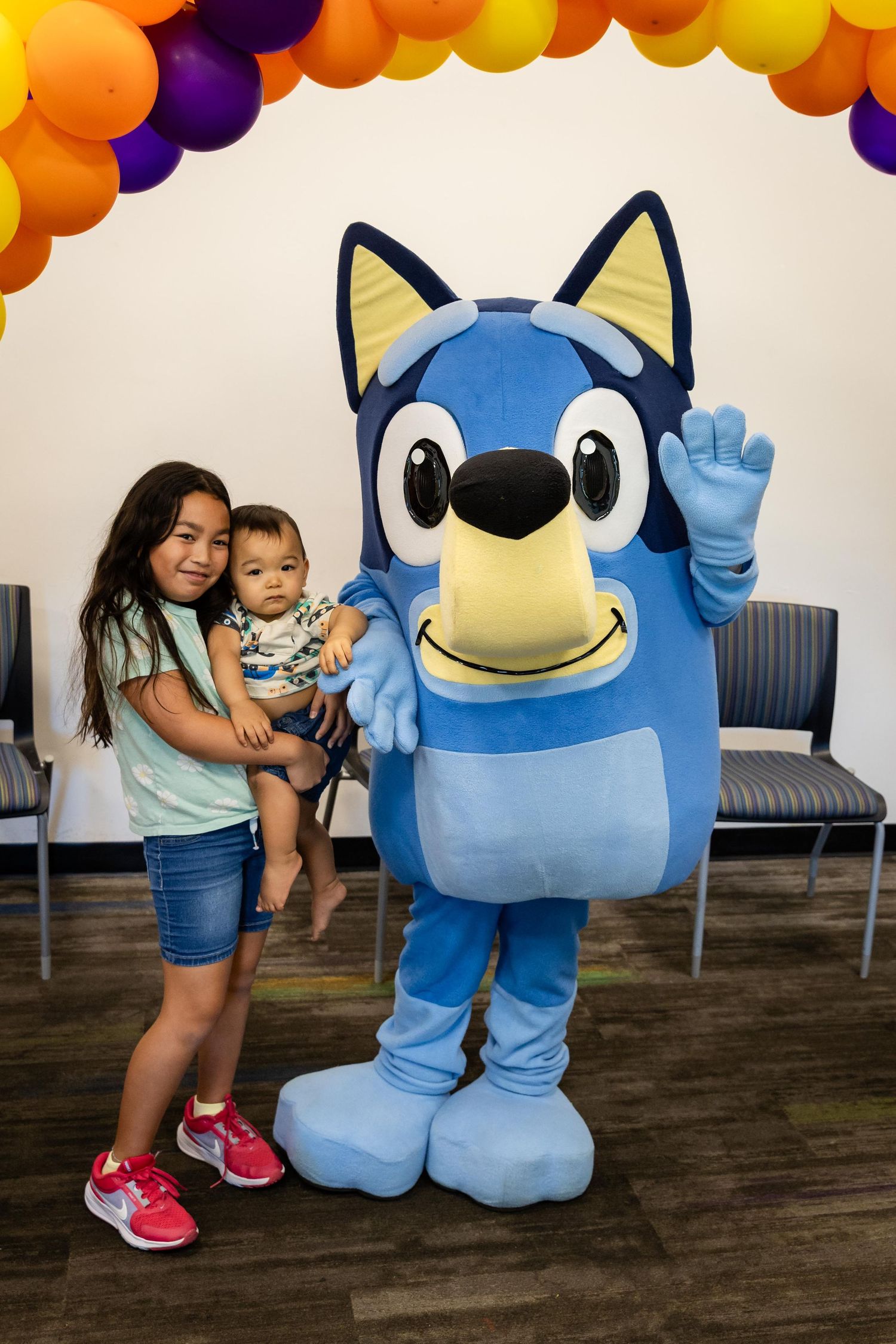 Girl holding baby poses with Bluey cartoon character mascot at colorful balloon arch party event.