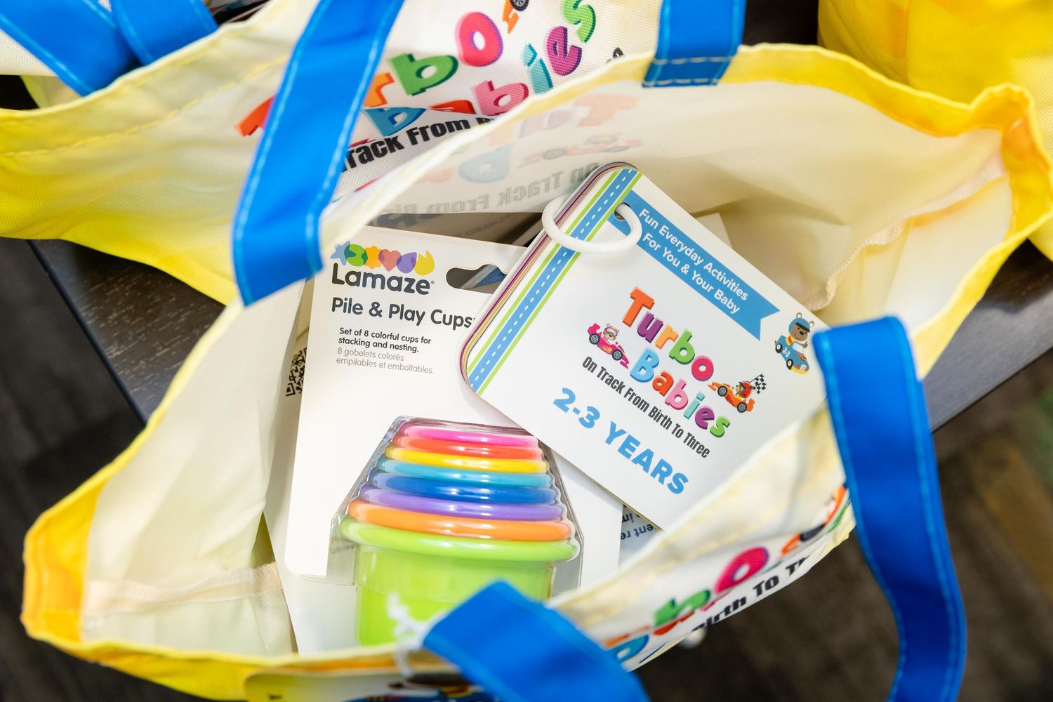 Yellow tote bag filled with Lamaze Pile and Play Cups and Turtles & Tails baby toys for ages 2-3 years.