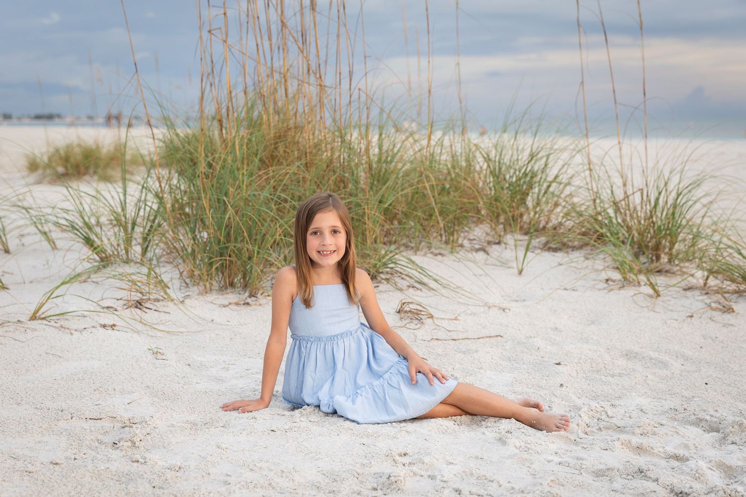 A young girl in a light blue dress sits smiling on white sand beach with sea oats in the background.