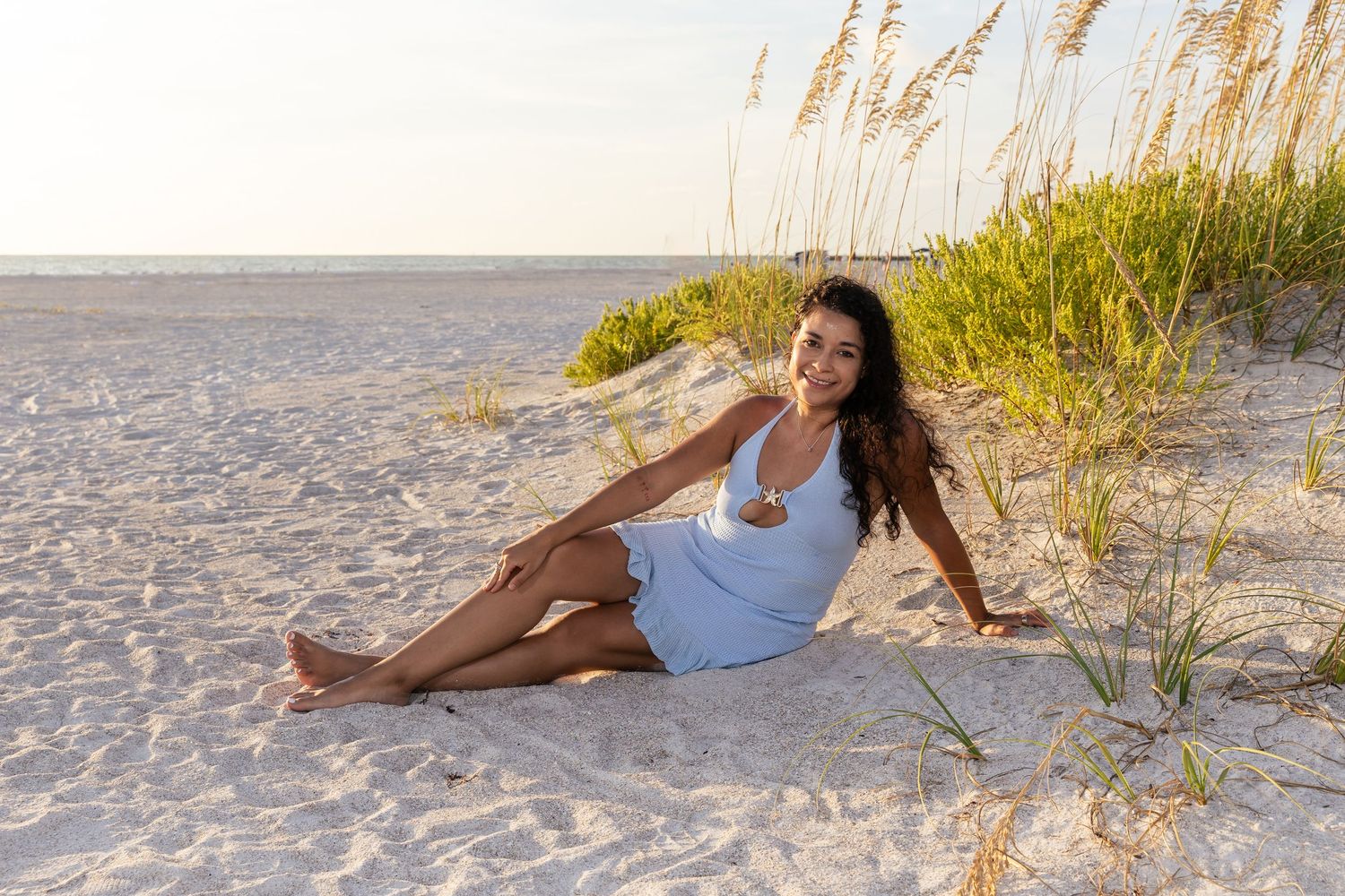 Young woman in blue dress sitting on sandy beach dunes with sea oats during golden hour.
