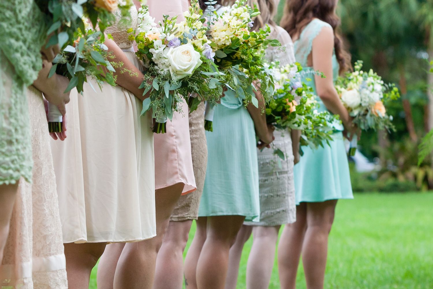 Row of bridesmaids in mismatched pastel dresses holding white and greenery bouquets standing on green grass Museum of Fine Arts, St. Peters.