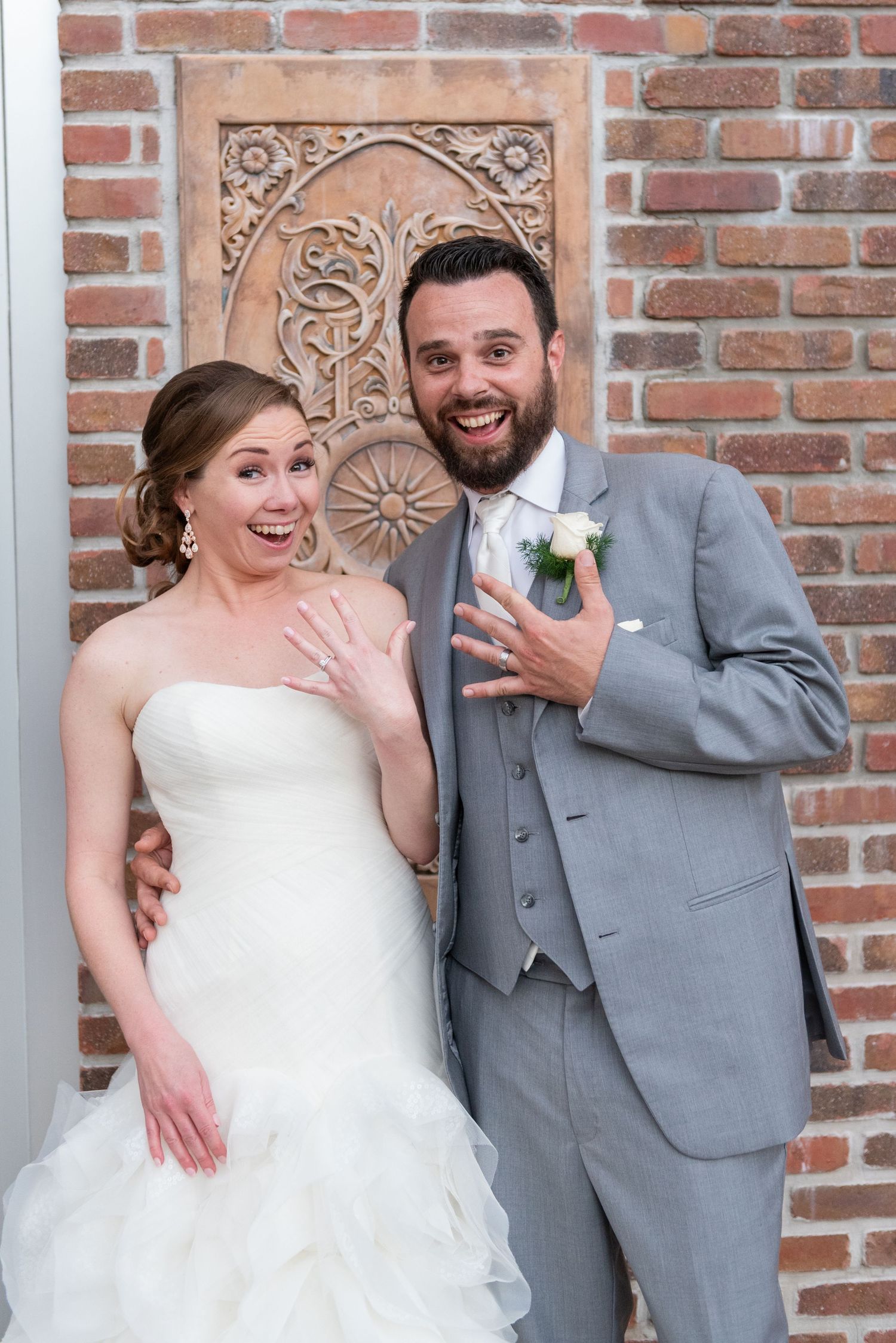 Joyful bride and bearded groom show off wedding rings against decorative brick wall backdrop.