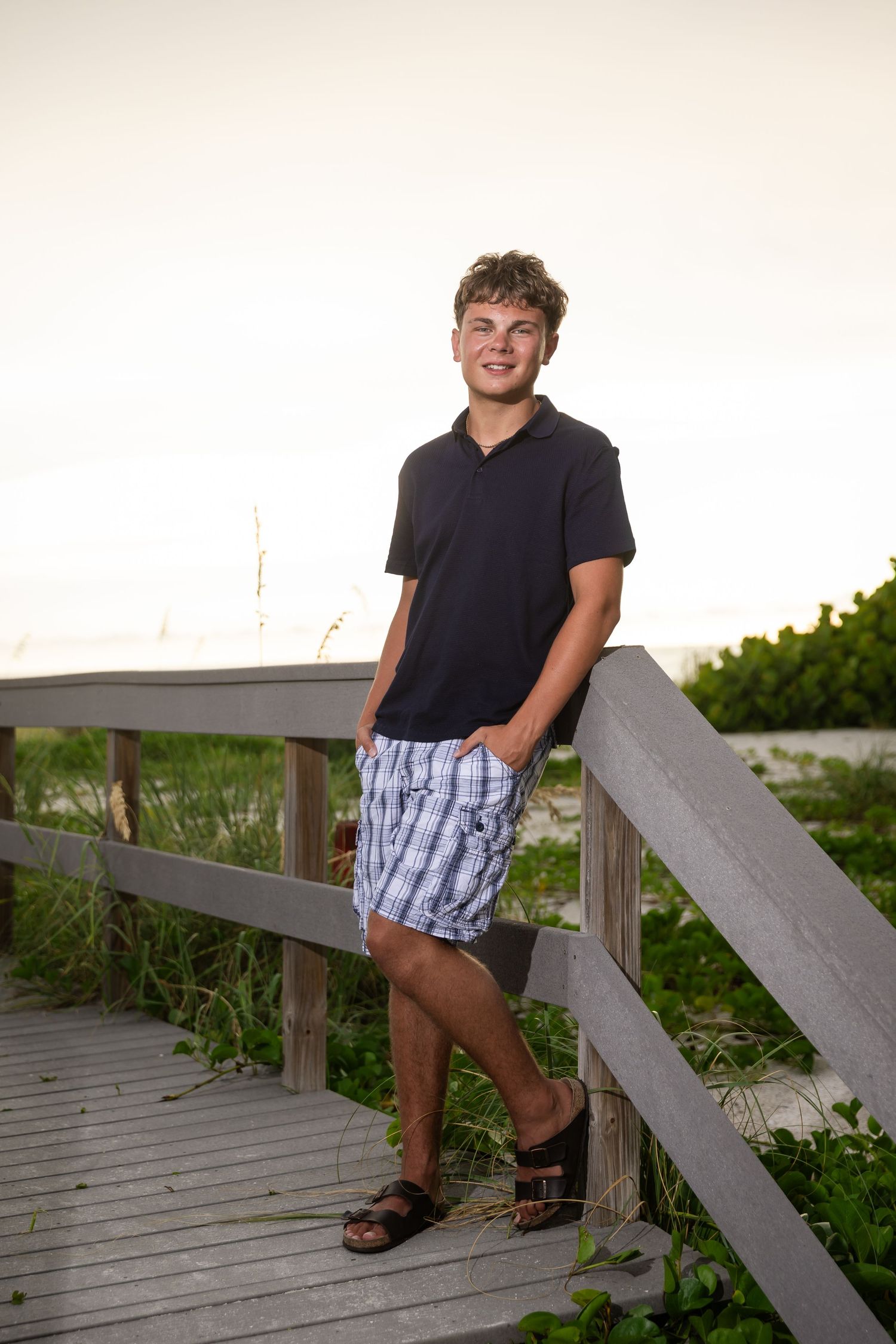 Young man in black polo and plaid shorts leaning on wooden boardwalk railing near beach vegetation at sunset.