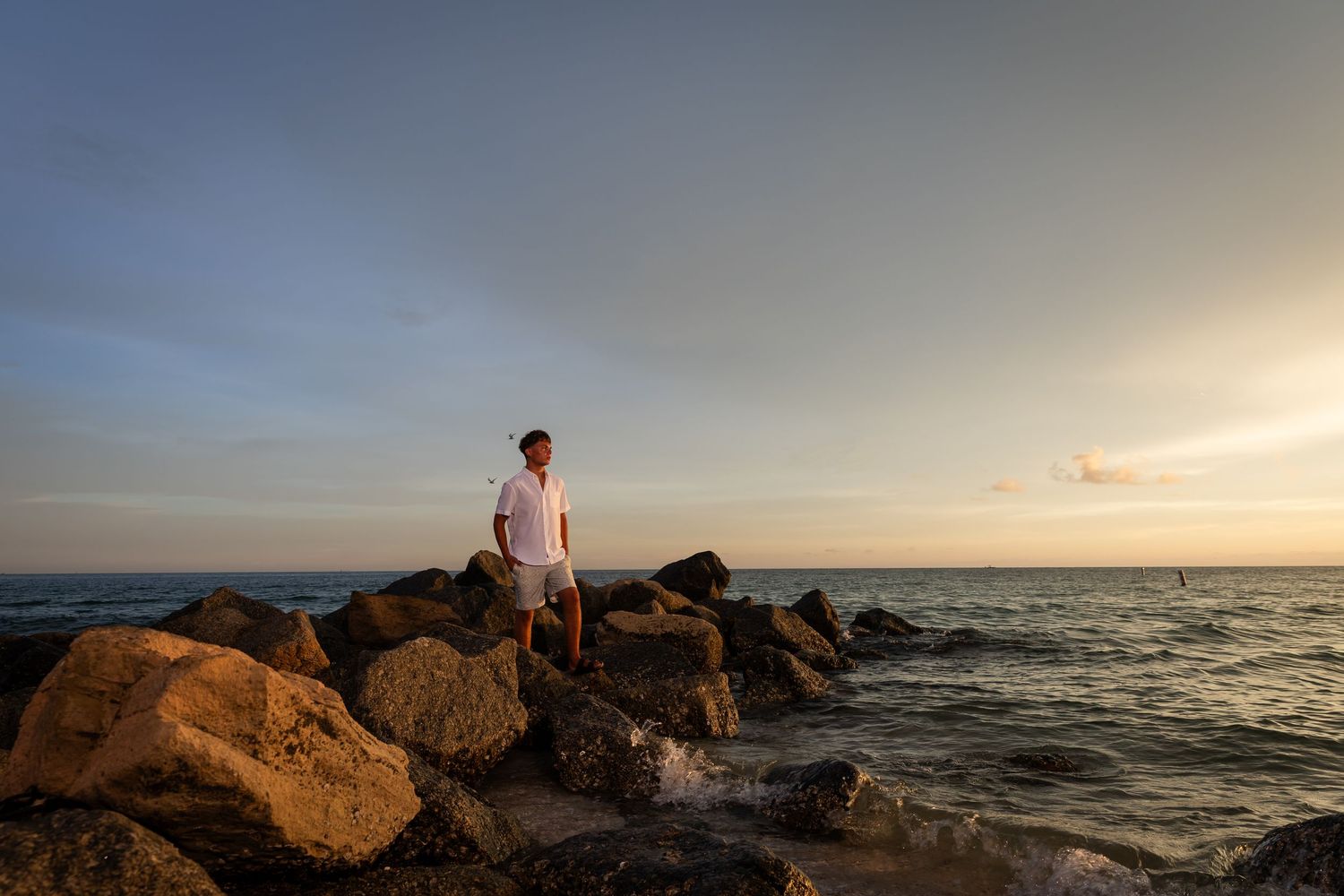A man stands on rocky shoreline at sunset, gazing at the ocean with warm golden light reflecting on the water.