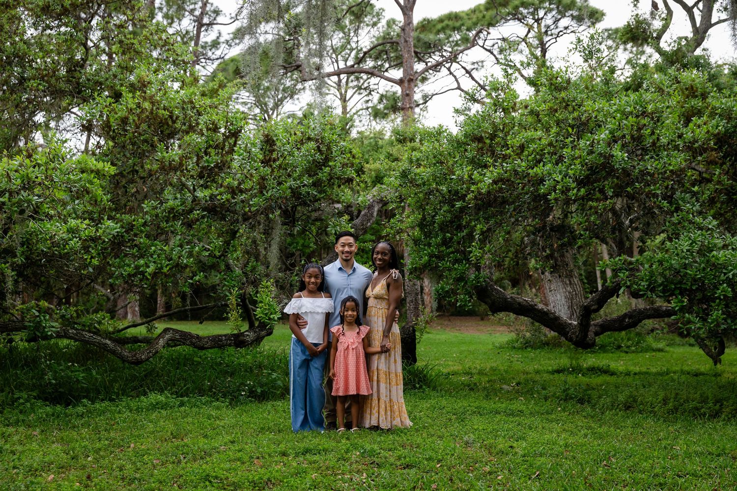 Family of four poses together on lush green grass surrounded by large oak trees in a scenic outdoor setting.