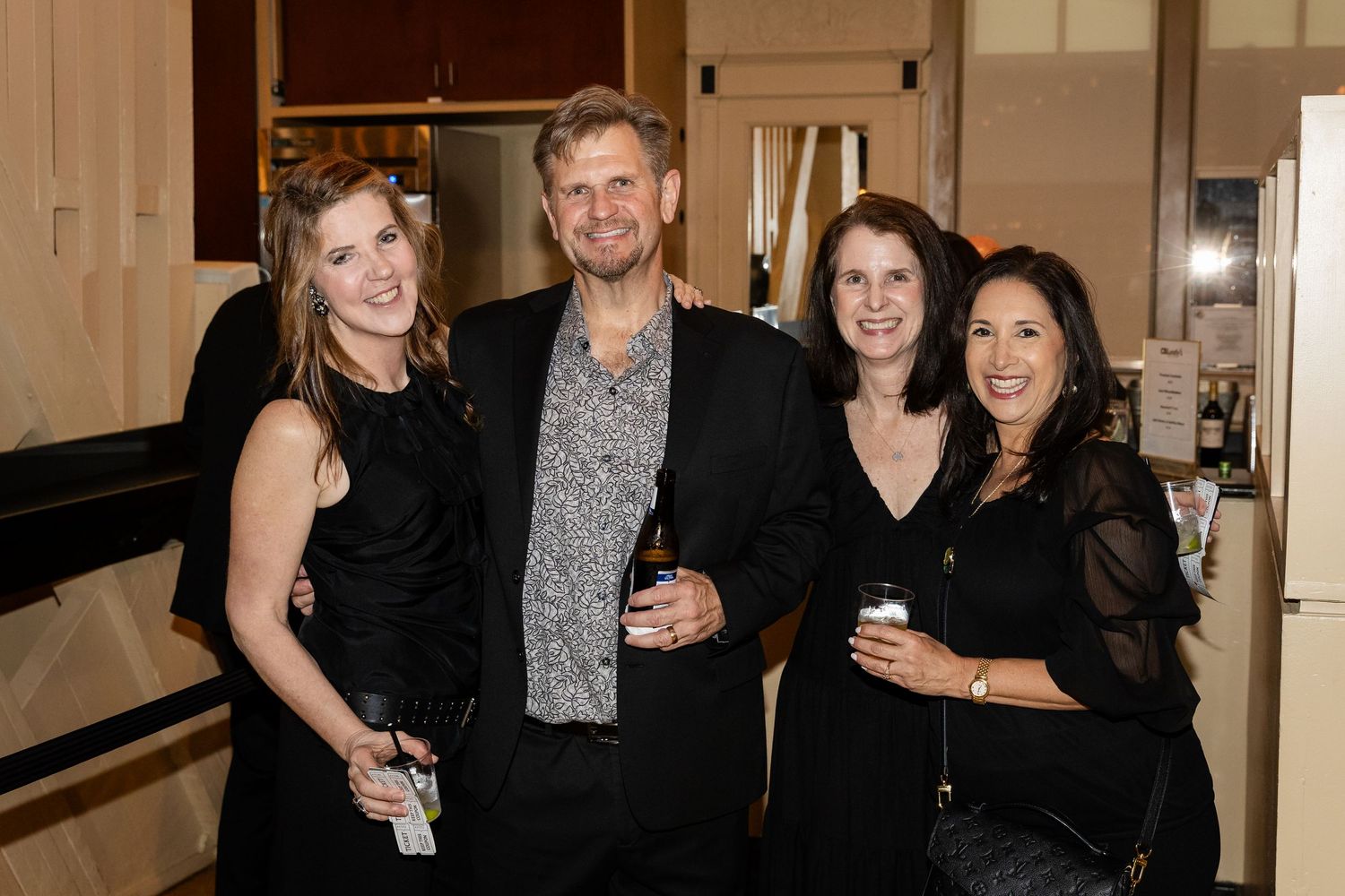 Four people smiling and posing together at a formal evening event, holding drinks and dressed in elegant black attire.