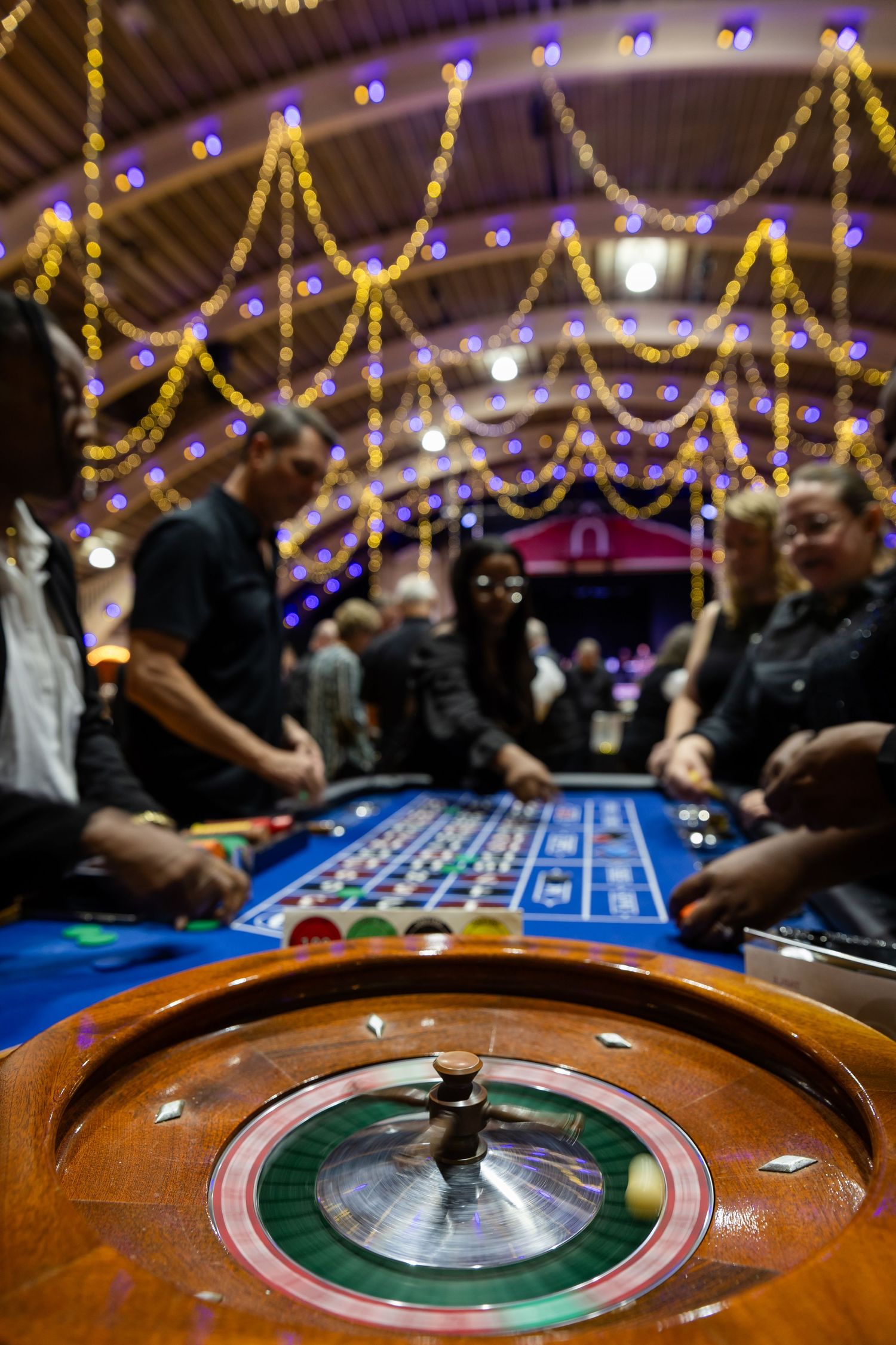 Close-up of a roulette wheel spinning at a casino night event with guests and festive lights in background.