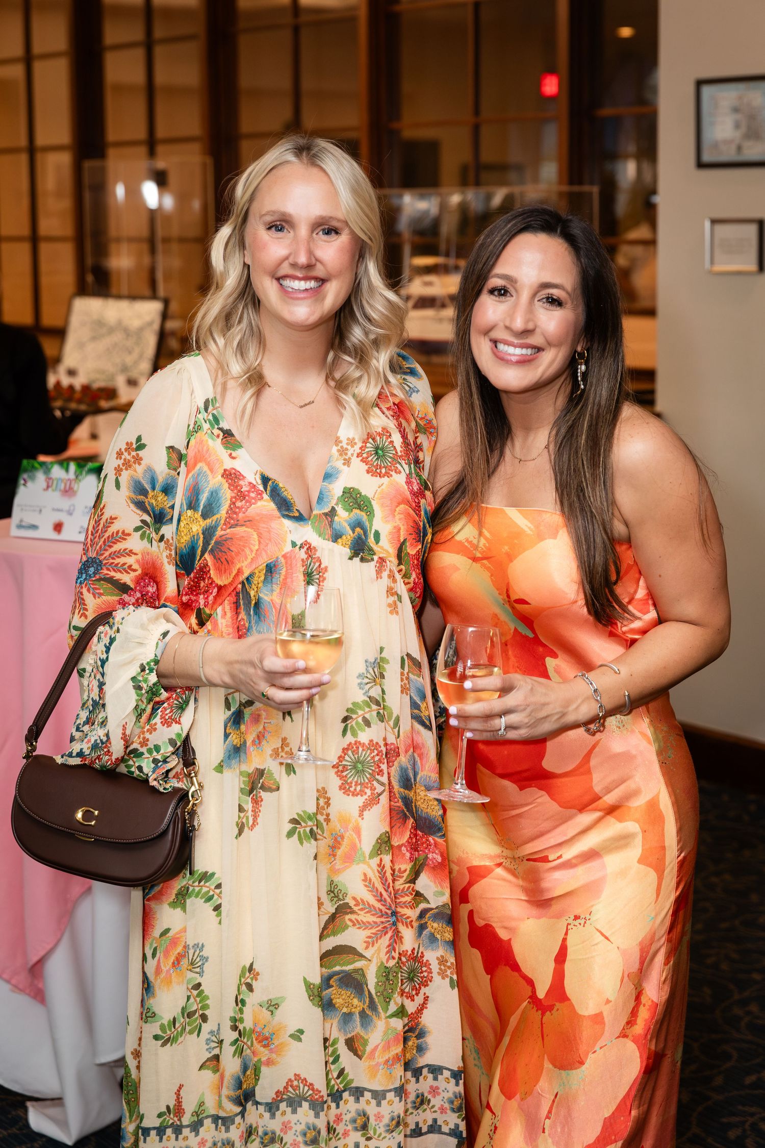 Two women in floral dresses smile and hold wine glasses at an elegant indoor event.
