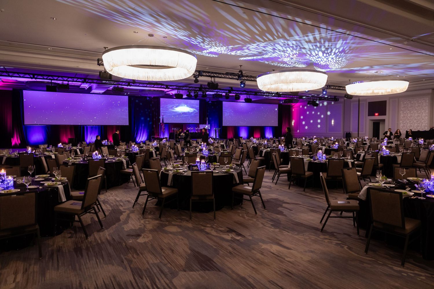 Elegant ballroom event setup with round tables, purple uplighting, and large circular chandeliers for a formal gala.