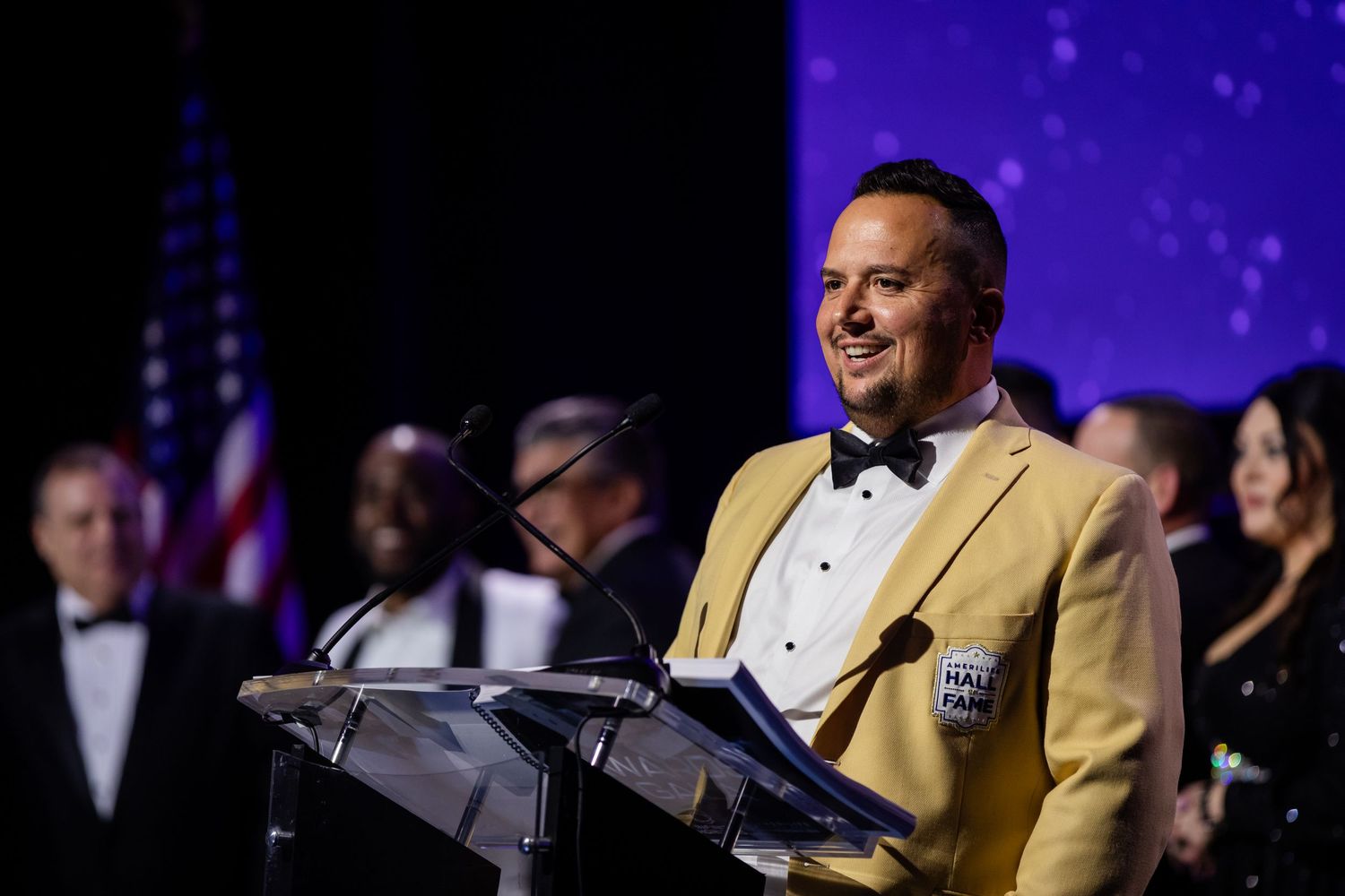 Man in gold blazer speaks at podium during formal Hall of Fame ceremony with American flag in background.