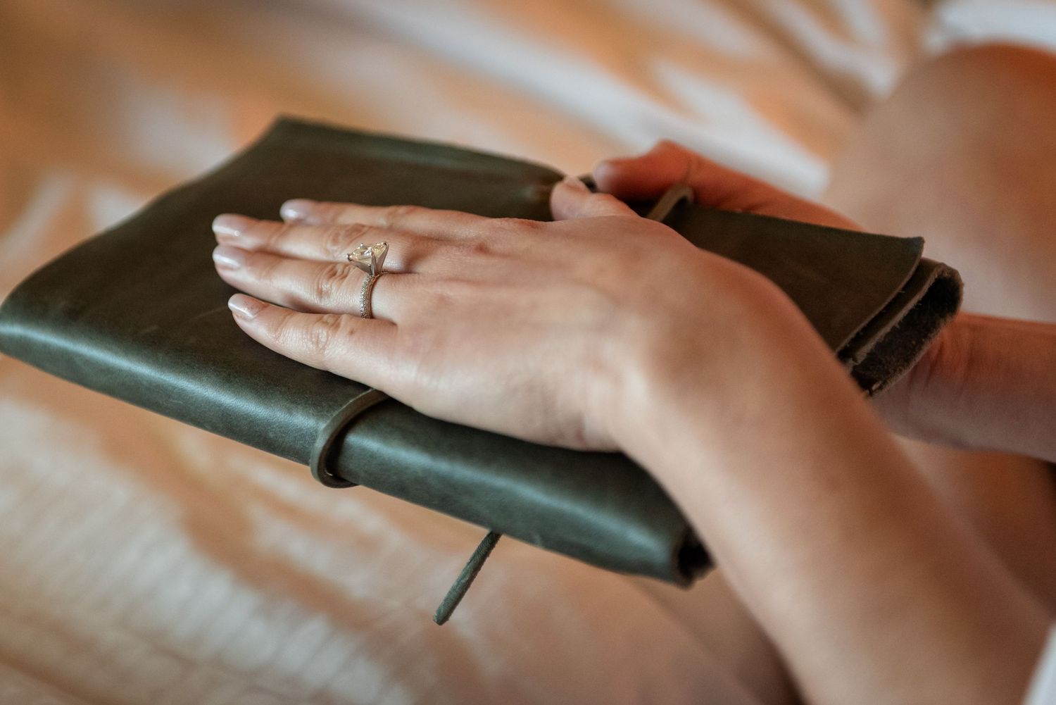 Hands with rings resting on a dark green leather journal, placed on a soft beige surface.