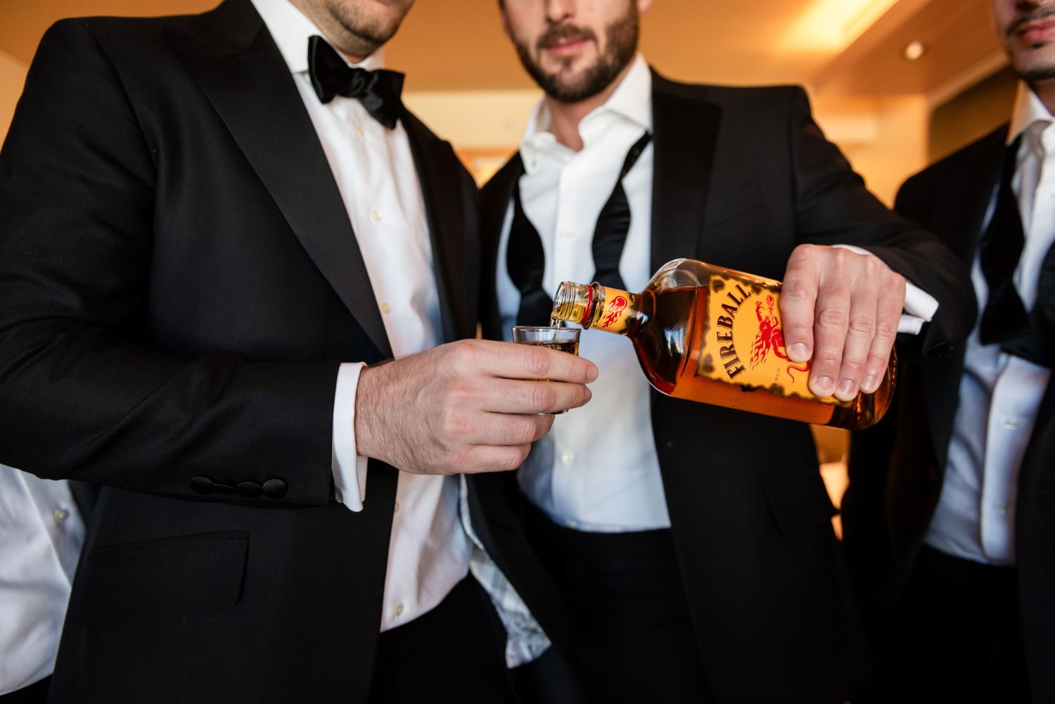 Groomsmen in black tuxedos pouring Fireball whiskey into a glass during wedding celebration.