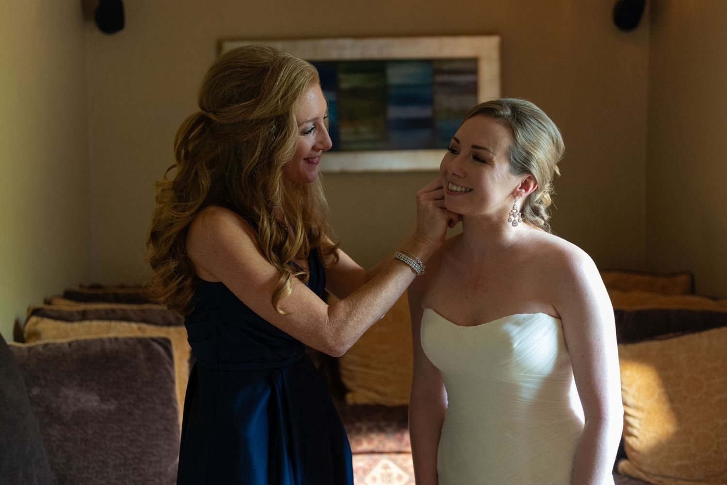 Bride in strapless white gown smiles as woman in navy dress gently touches her face during wedding preparations.