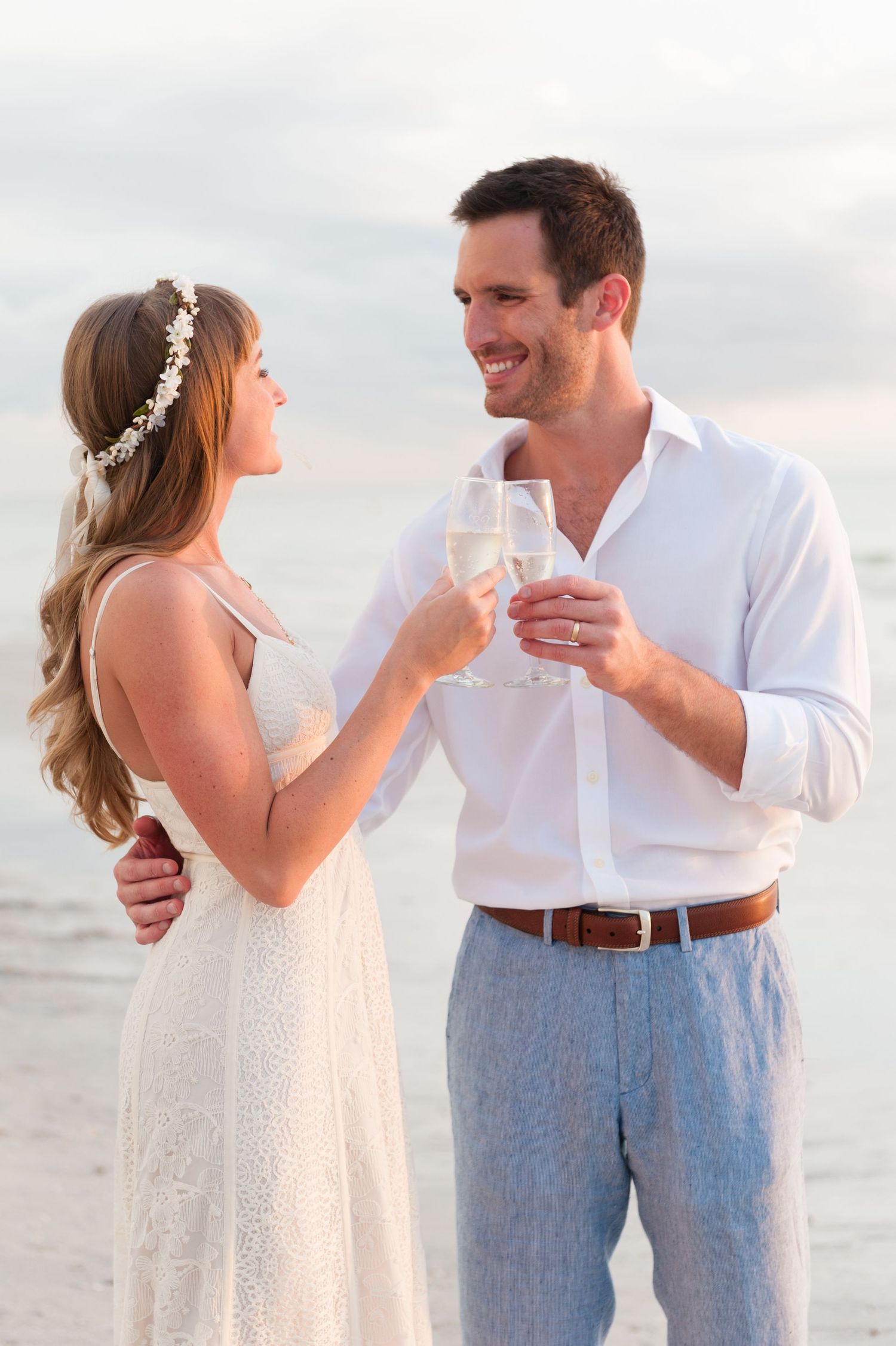 Couple toasting champagne glasses on beach at sunset, bride in white lace dress with floral crown, groom in white shirt.