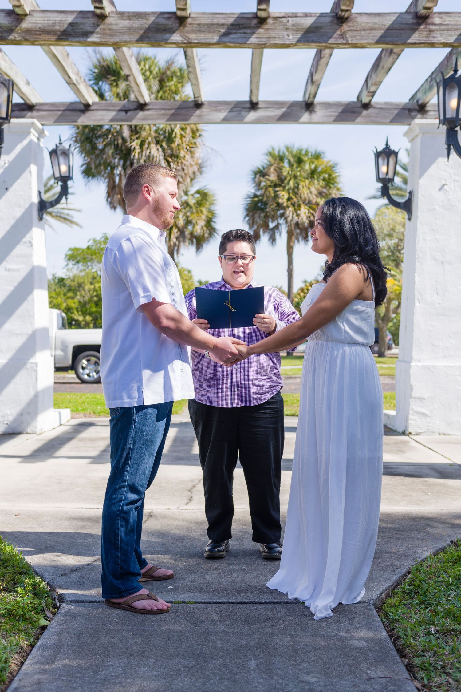 Couple exchanges vows outdoors under a pergola with an officiant holding a book, surrounded by palm trees on a sunny day.