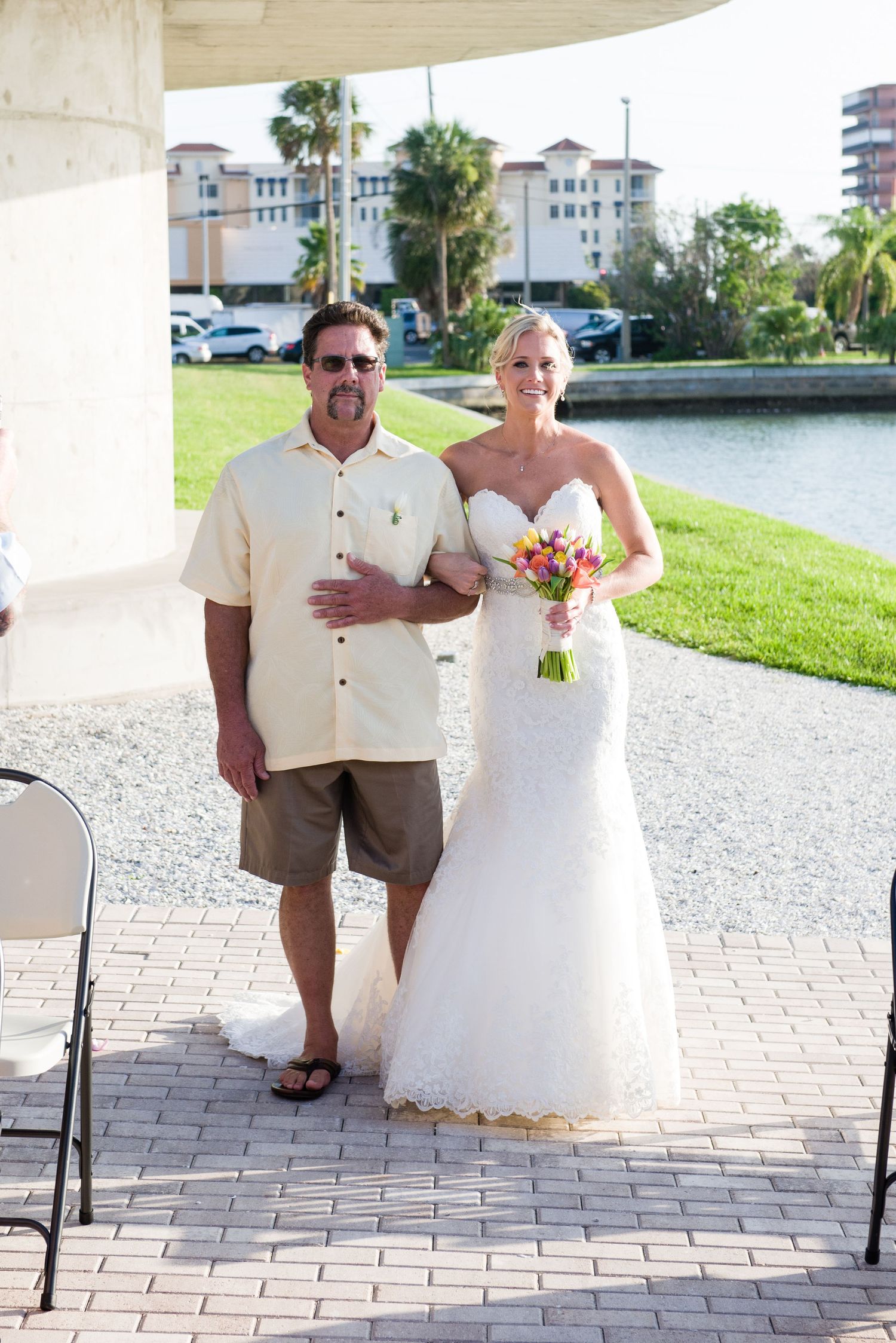 Bride in white gown holding colorful bouquet walks with man in casual attire at outdoor waterfront wedding ceremony.