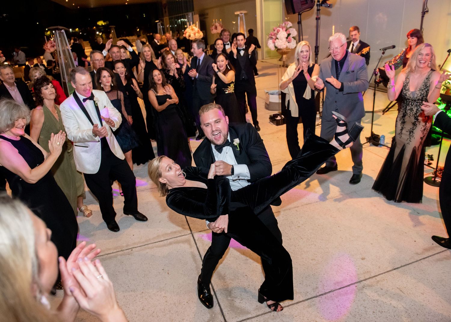 Groom dramatically dips bride on dance floor surrounded by cheering wedding guests during reception.