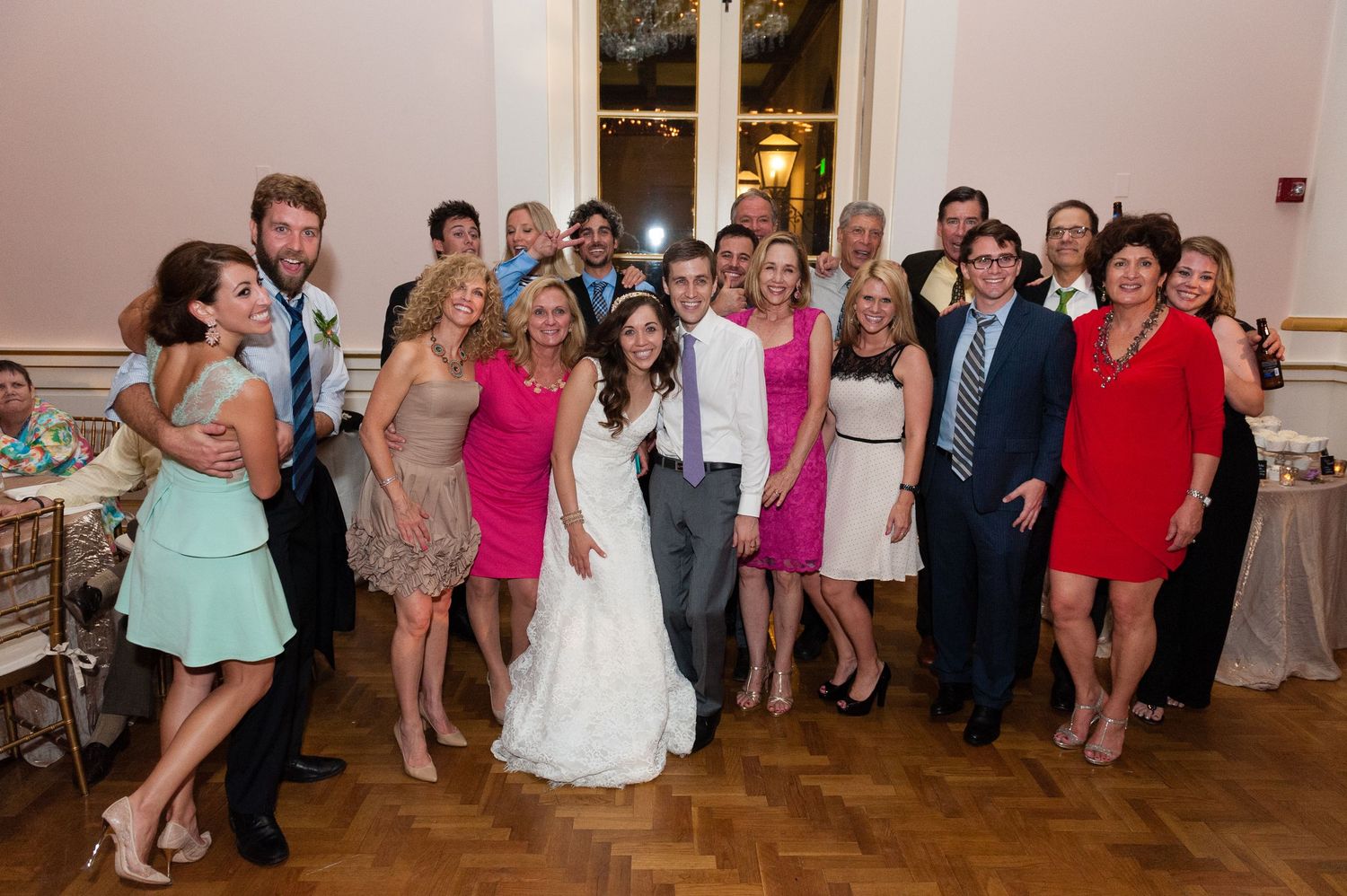 Wedding party group photo with bride in white gown surrounded by guests in colorful dresses on dance floor.