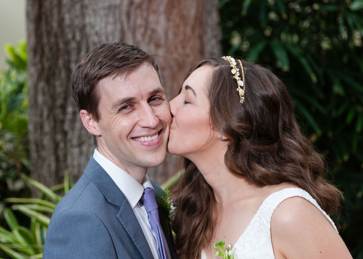 Bride in white lace gown kissing groom on cheek while he smiles outdoors near greenery.