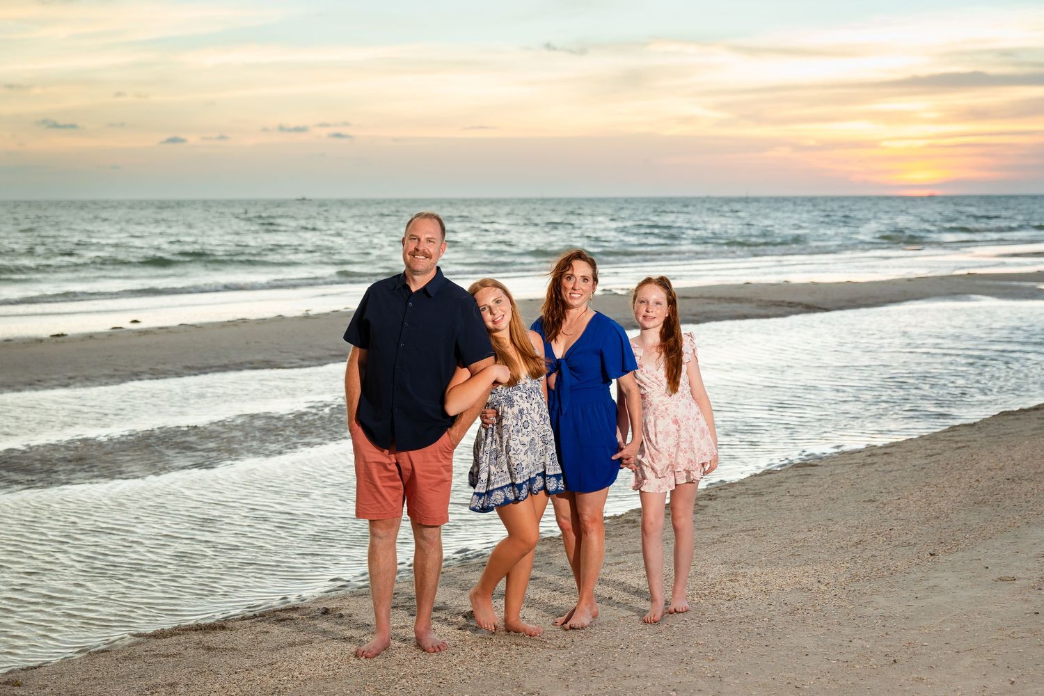 Family of four posing on a sandy beach at sunset with colorful sky and ocean waves in the background.