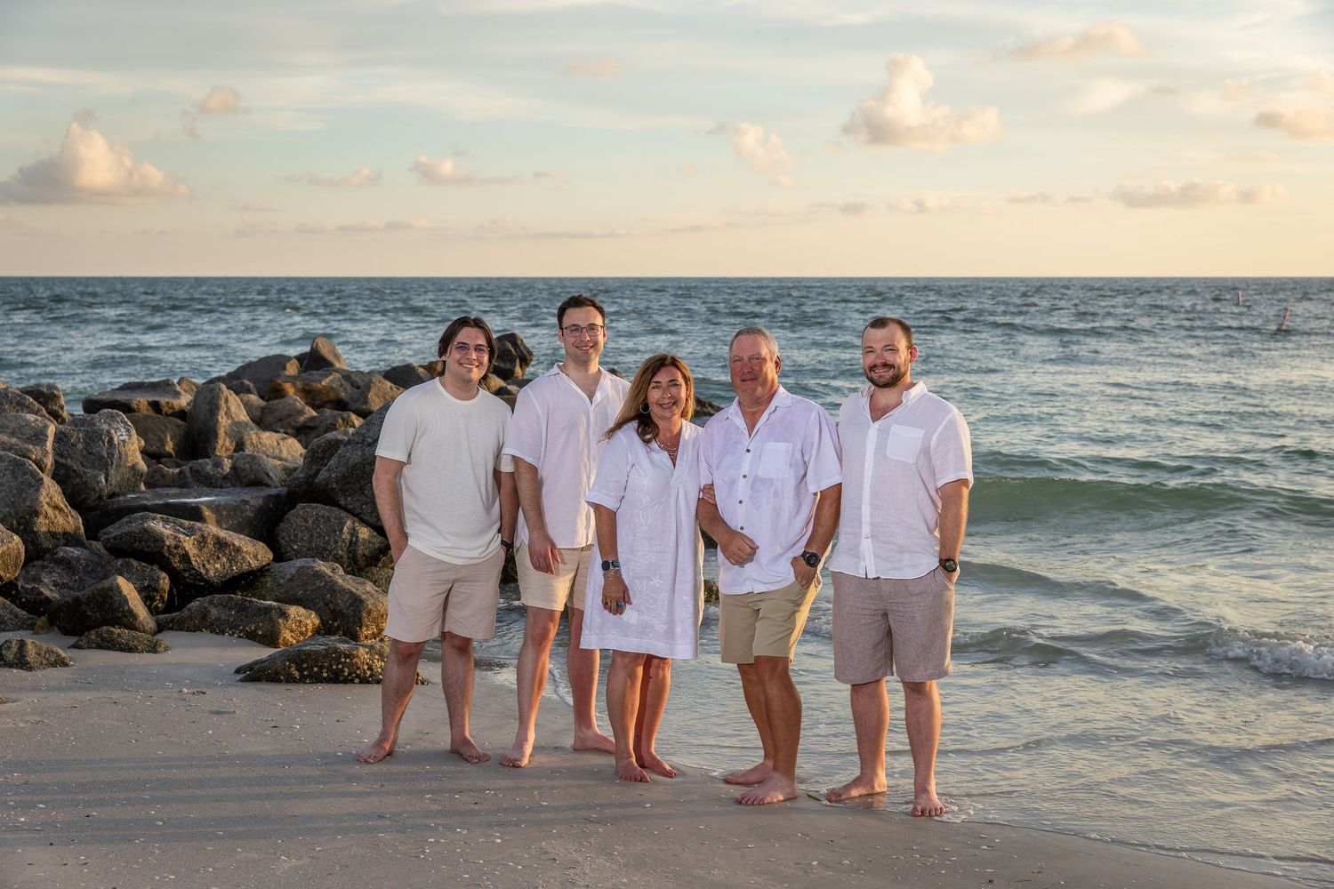 Family of five posing together on a sandy beach at sunset near rocky jetty, wearing white and khaki outfits.