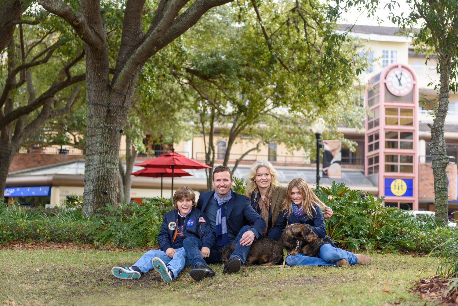 Family of four with two brown dogs sitting on grass under trees in a park setting with a clock tower in the background.